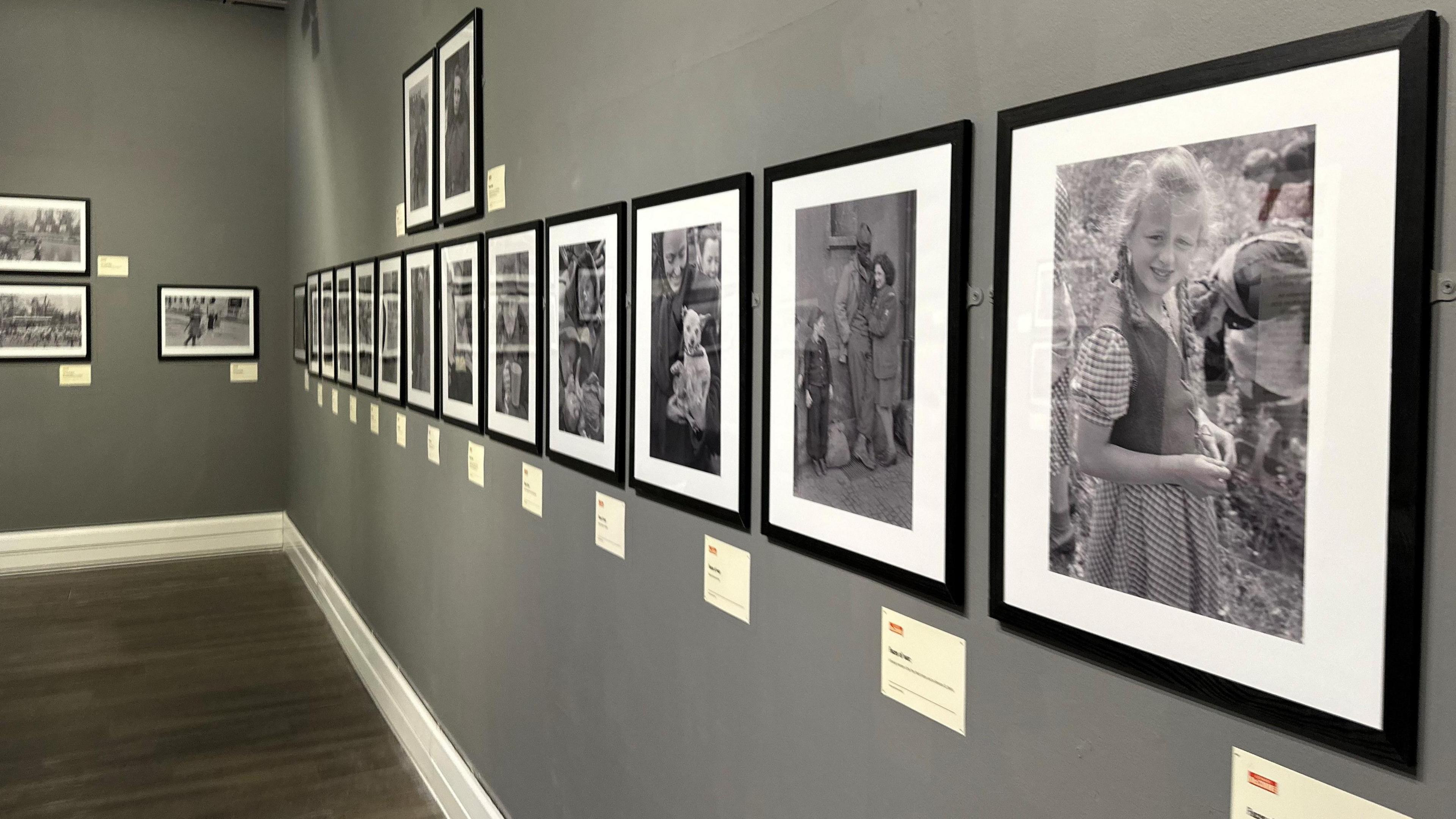 A row of photographs in black frames hanging on a grey gallery wall, The one at the front features a fair-haired child with plaits smiling.