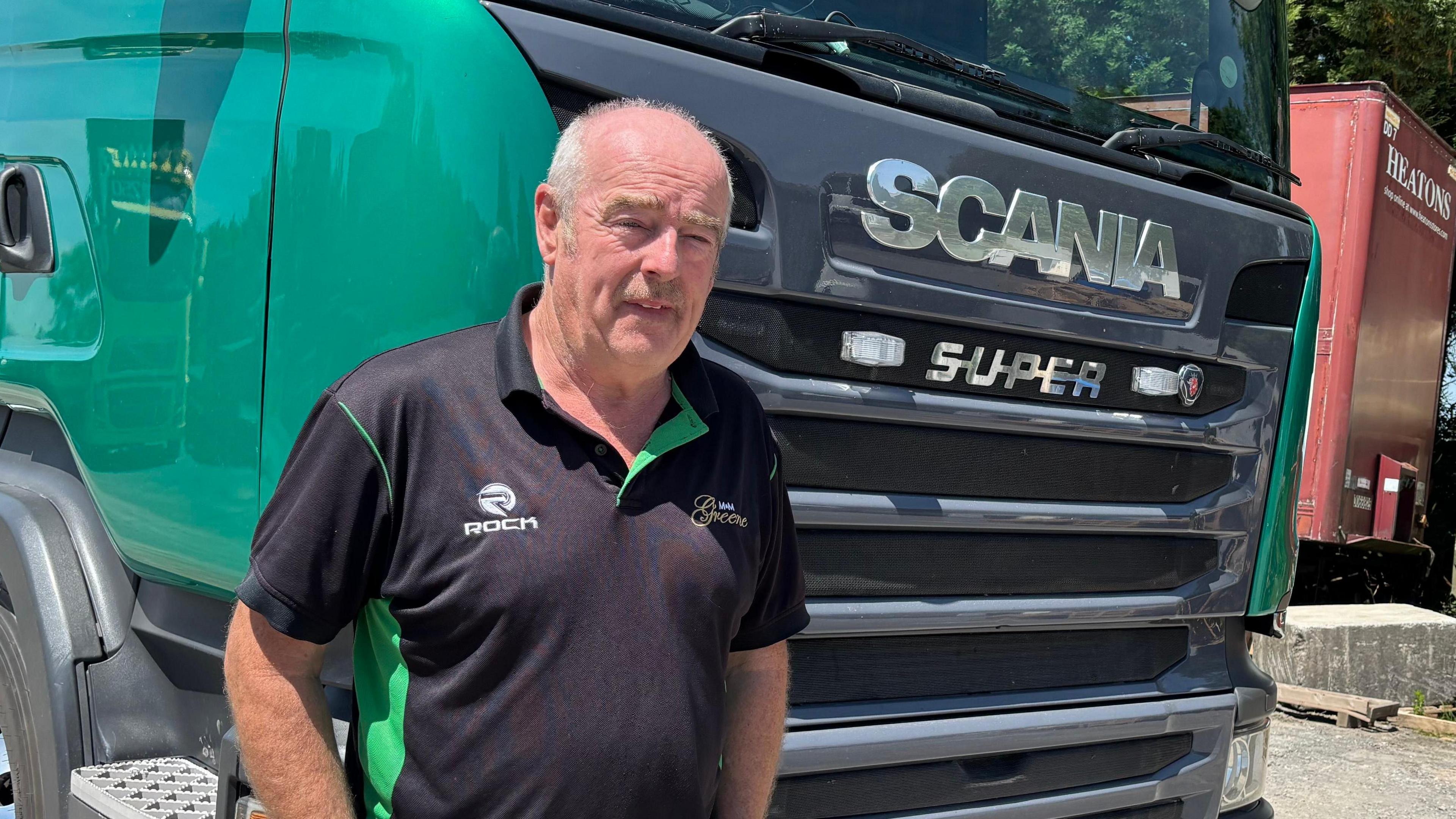 Mike Greene mewn crys polo du yn edrych ar y camera, yn sefyll o flaen cab lori anferth gwyrdd a du
