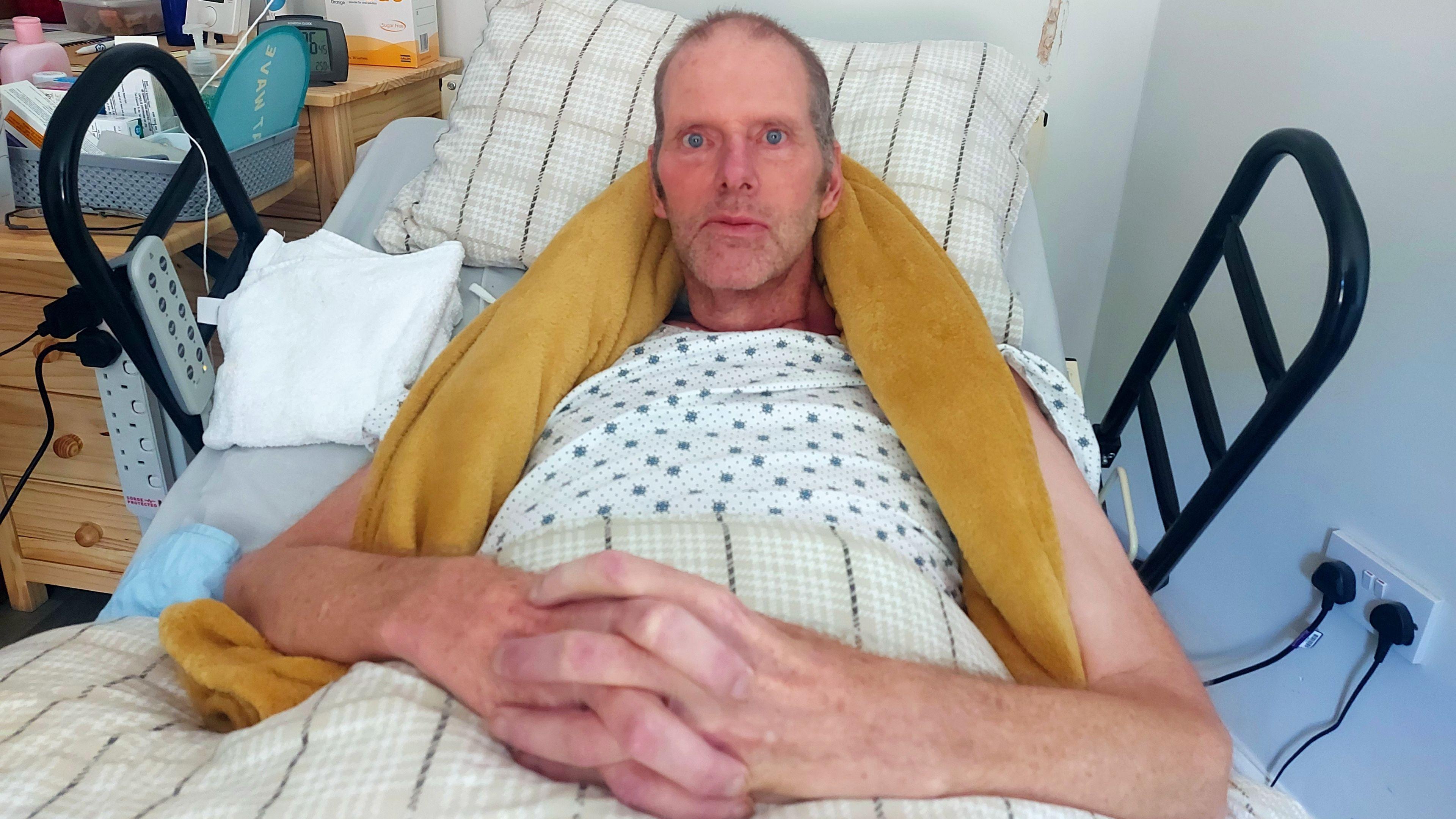 Tim Hull lying in an NHS hospital bed in his home. He is clasping his hands together and looking towards the camera. He has a yellow towel around his neck for support and is wearing a white t-shirt with blue spots.