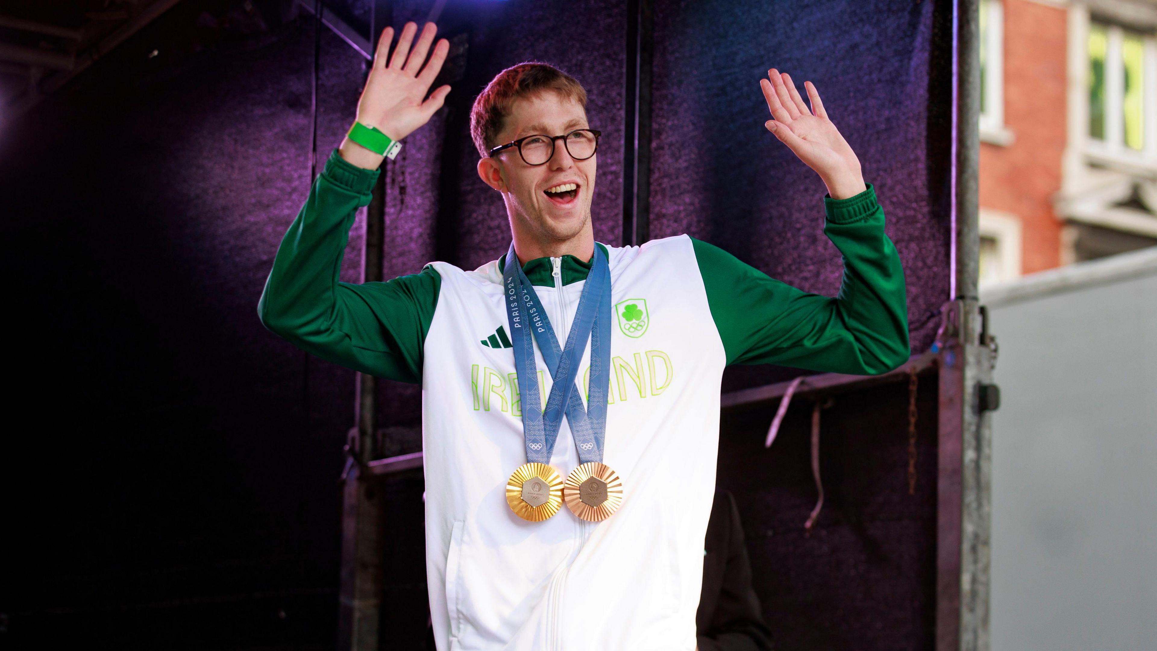 Daniel Wiffen, wearing his bronze and gold medals, waving both arms and smiling