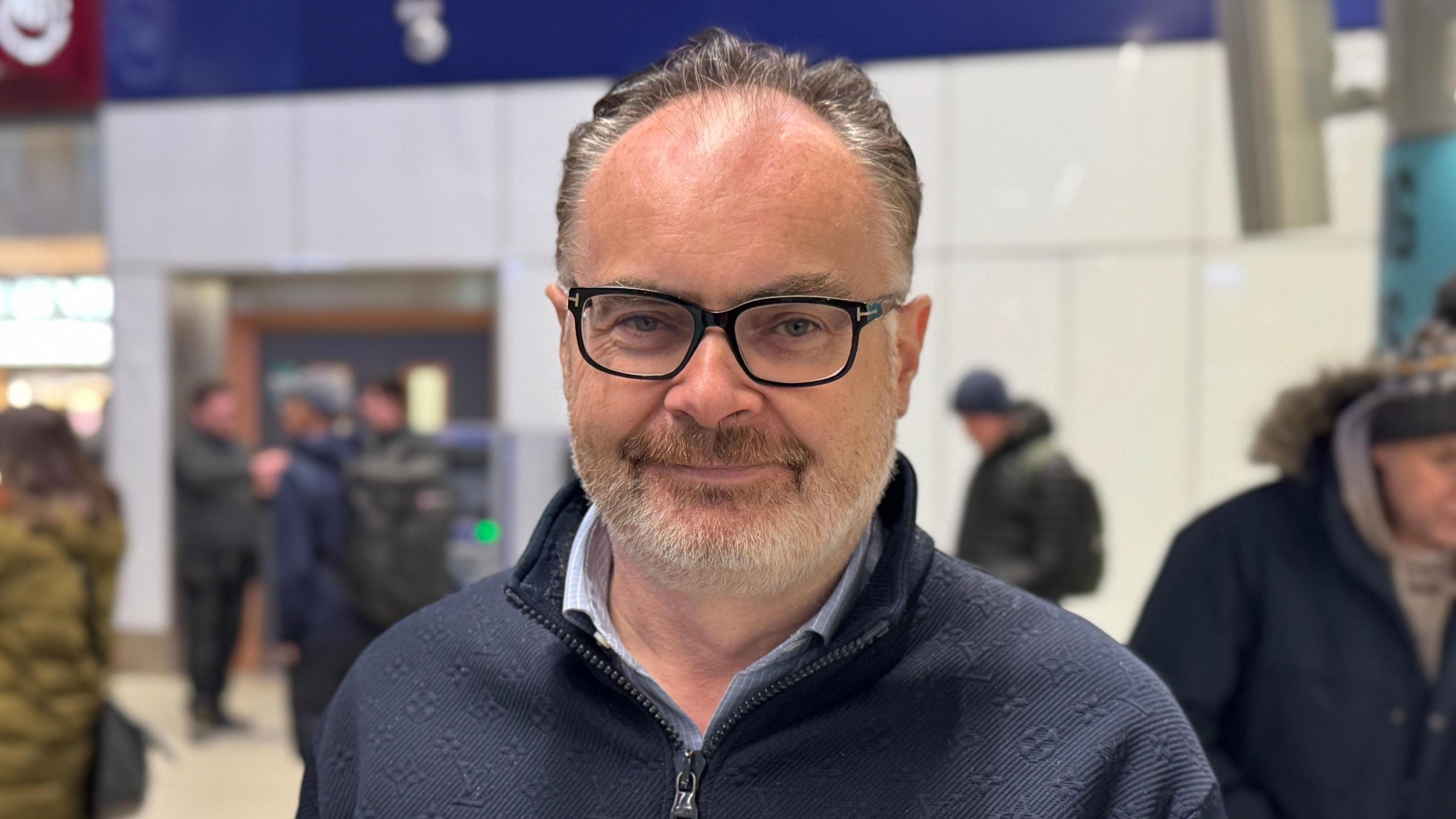 Troy Armour has short grey hair and a white beard. He is wearing a navy zip up jumper and a light blue shirt underneath. He is also wearing black framed glasses. 