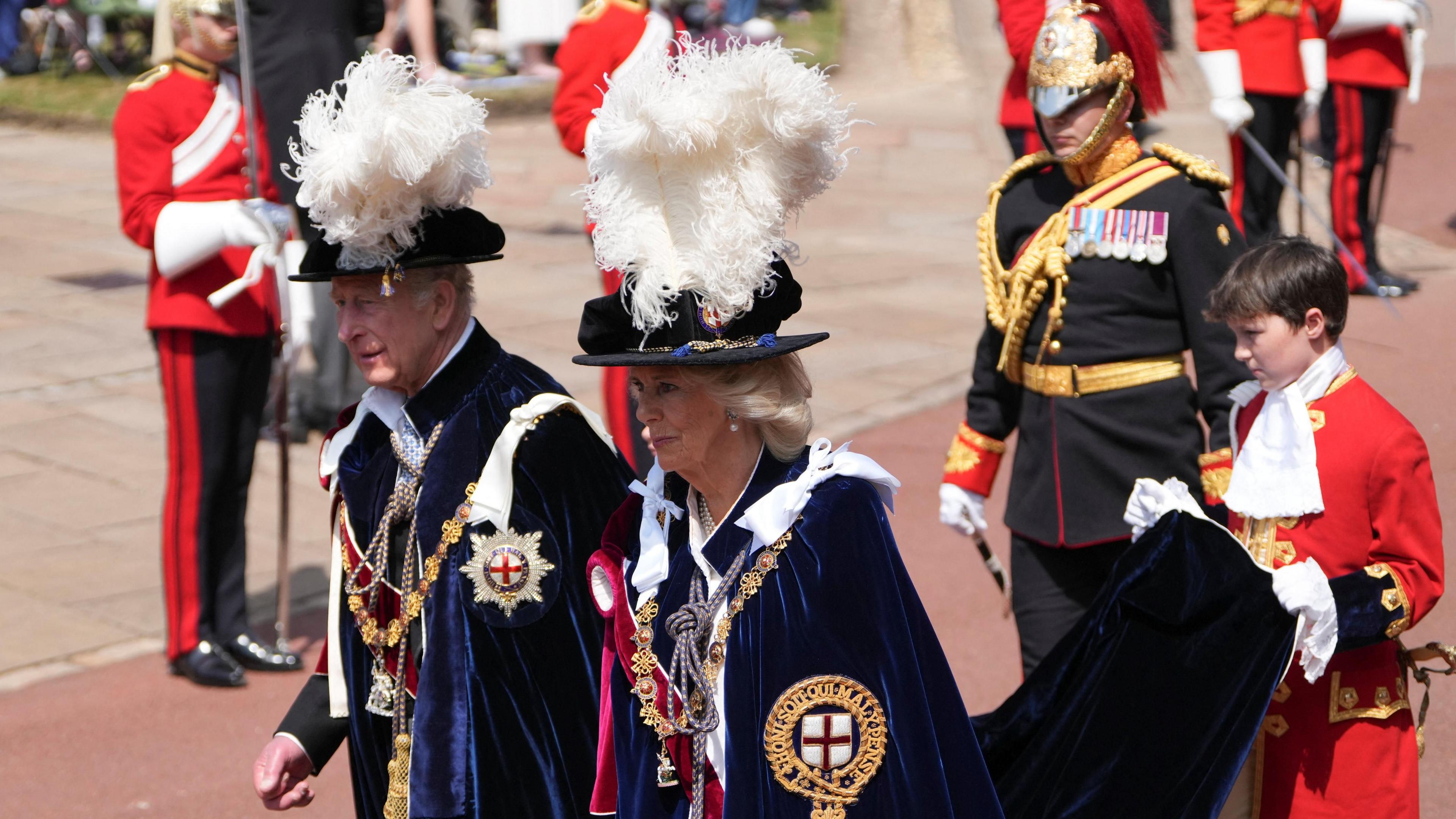 King Charles and Queen Camilla arrive for the Order of the Garter service in traditional robes. A boy in red uniform holds up the end of the king's cloak. Guards with swords are behind.