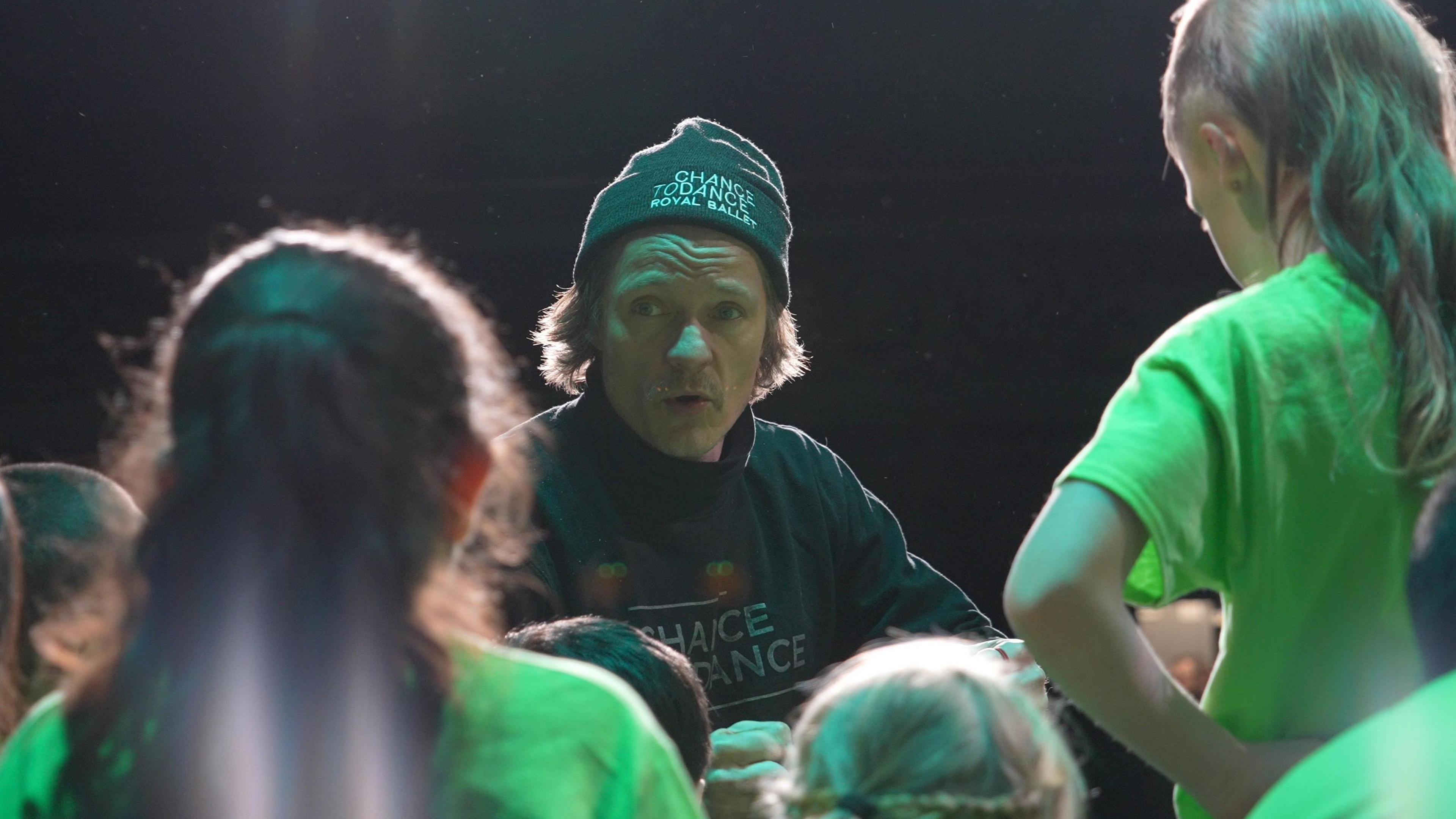 A man in a black hat is surrounded by children wearing green T-shirts on a stage.