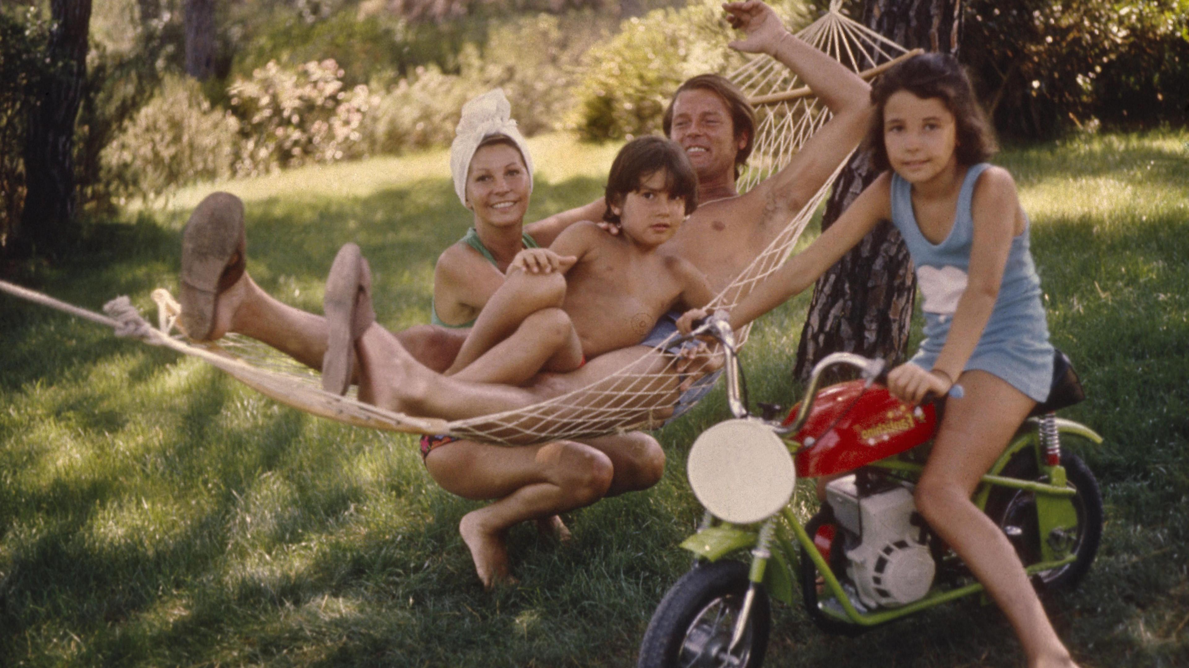 Sir Roger Moore lies on a hammock. He is wearing swimming trunks and has his arm outstretched above him. He is smiling and looking relaxed. His wife Luisa is crouching down alongside the hammock. She is also wearing swimwear and has a towel wrapped around her hair. She looks content. Sir Roger's young son Geoffrey is sitting on his lap. He has dark brown hair and is looking at the camera apprehensively. Sir Roger's daughter Deborah is sitting on a small green motorcycle beside the hammock. She also has dark brown hair and is wearing a pale blue dress. She has a half smile on her face.