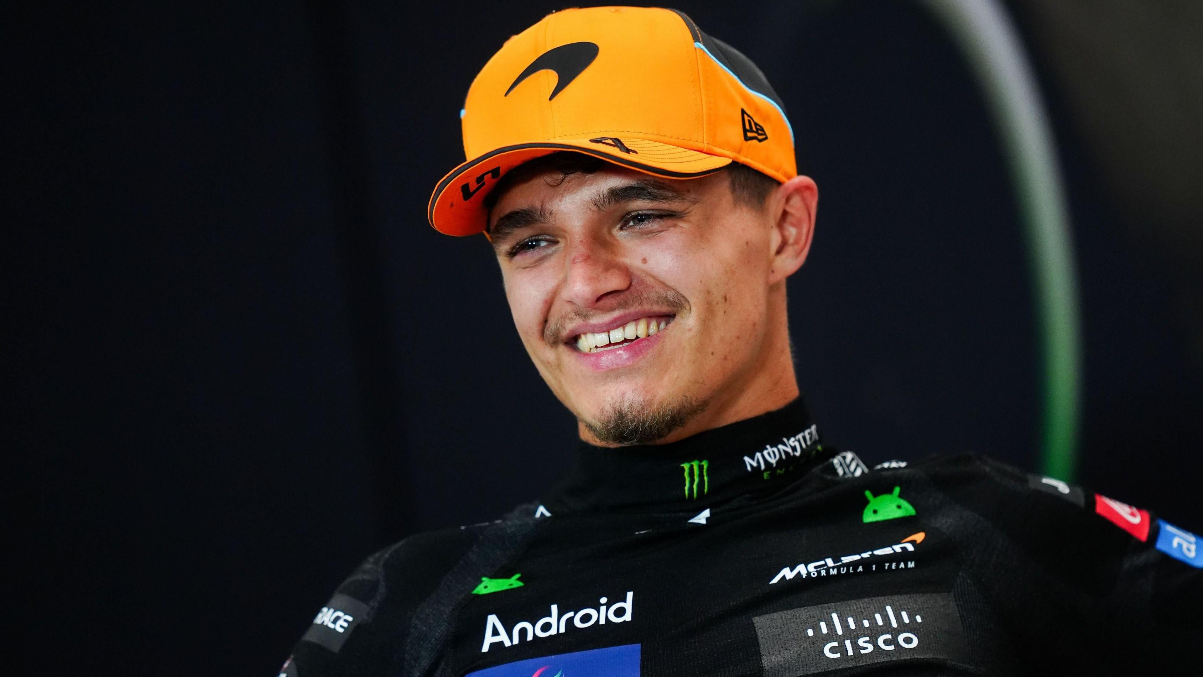 Image of Lando Norris. He is wearing a bright orange hat with the black McLaren logo. He is wearing a black top with various logos on it, including McLaren and Android. He is pictured looking to the side of the camera and smiling.
