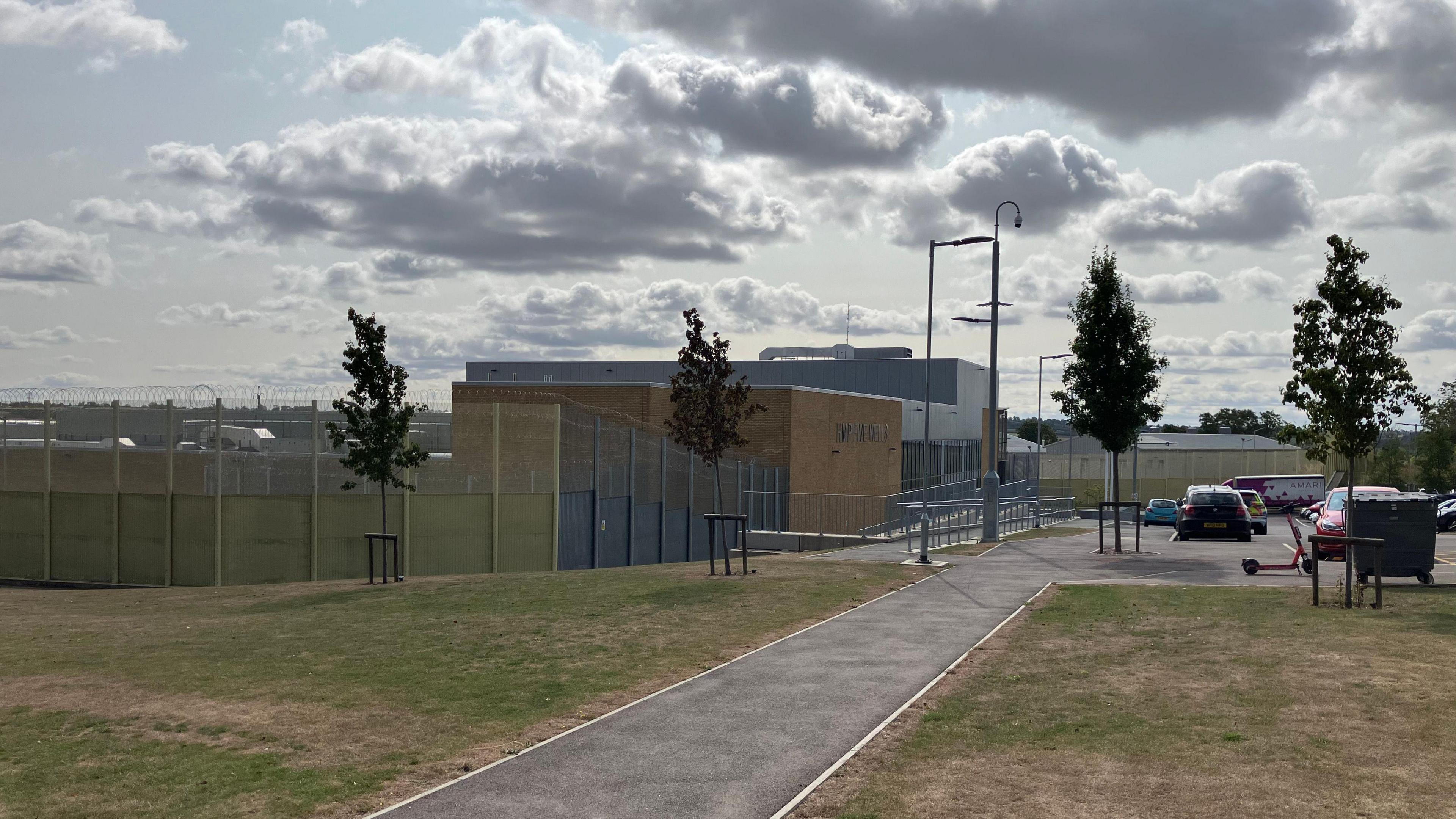 The high fences of a prison yard are shown from the outside, with buildings in the background and a car park, pavement and grassed area shown in the foreground.