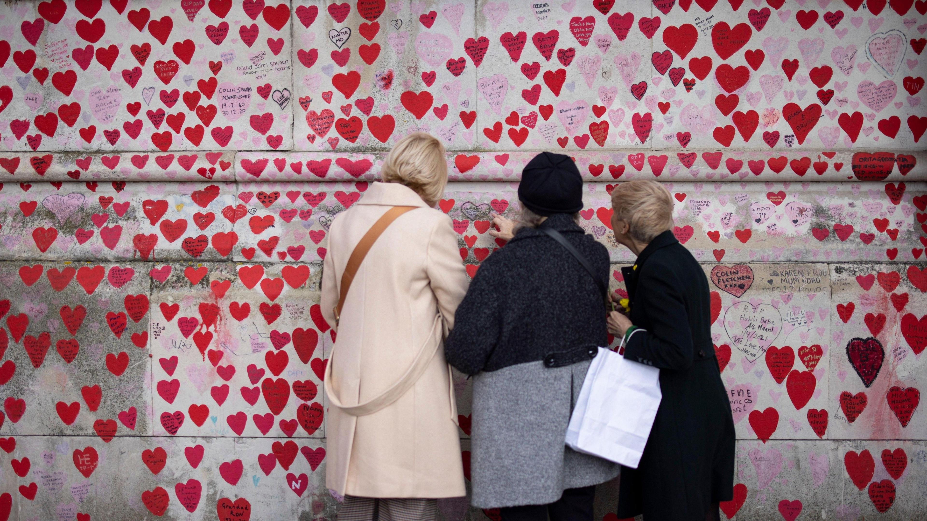 Three women inspect the hearts painted on the national Covid memorial wall. The red hearts can been on the grey of the stone with the backs of the three women facing the camera.