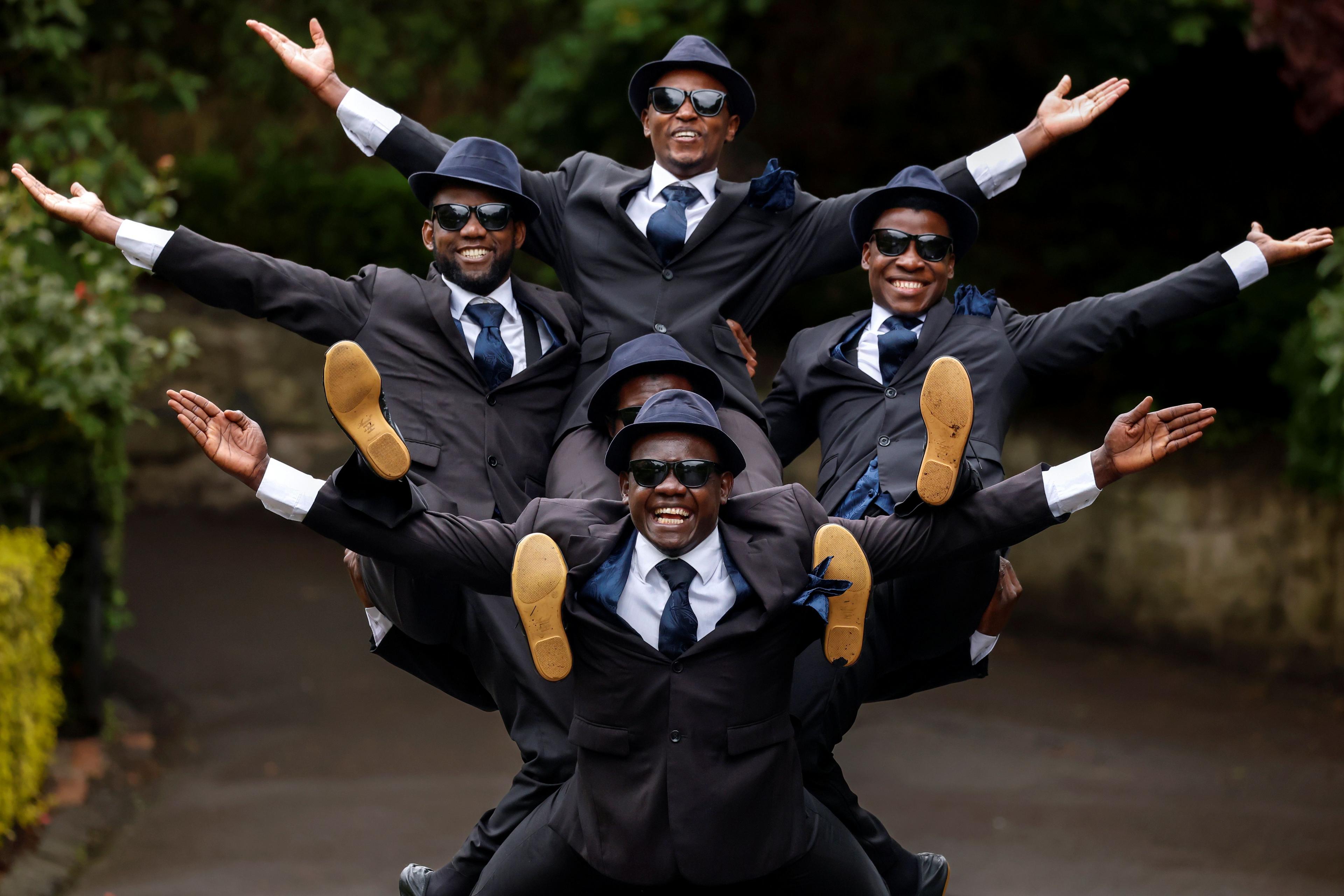 The Black Blues Brothers pose during a photocall for their Fringe show. Five men wearing matching dark suits, ties, hats and sunglasses are arranged in a stacked formation, with arms extended outward and legs raised.