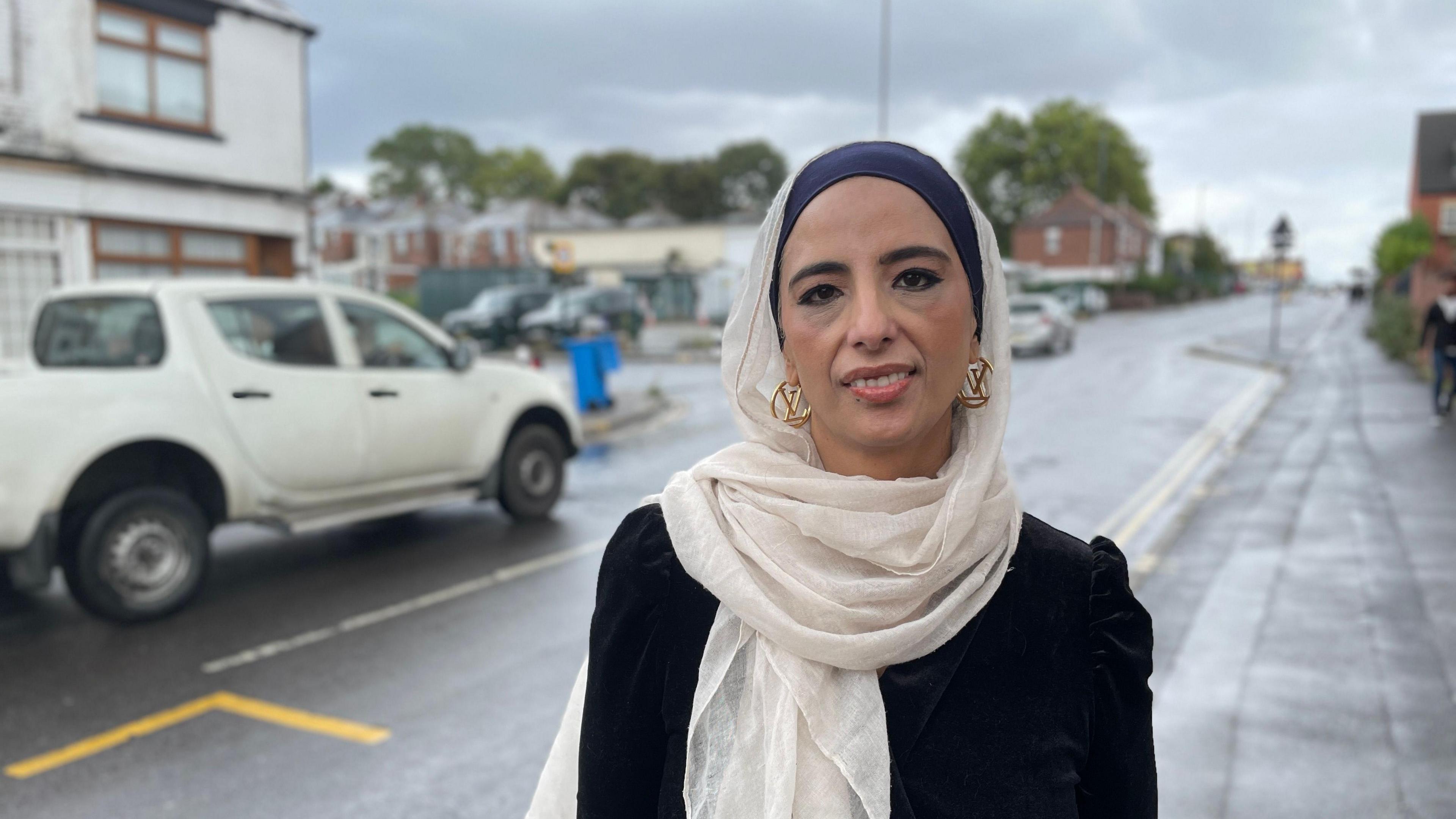 Campaigner Shama Abdaljawwad standing on the pavement of Staniforth Road in Sheffield. She's smiling and is wearing gold earrings and a cream scarf.