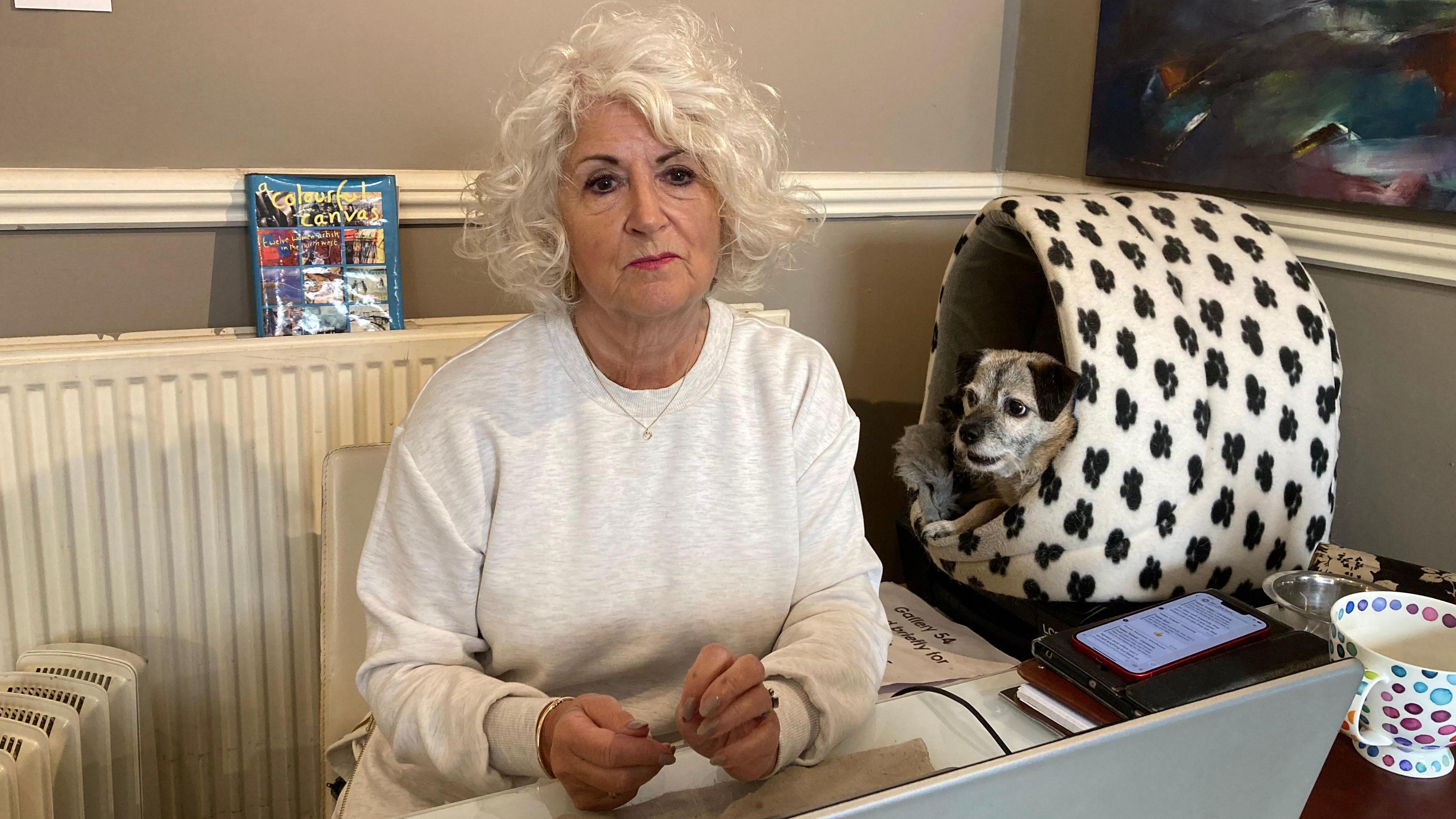 A woman with long white hair sits at  a desk with a small dog in a white dog bed with black paw prints, placed on the table next to her. A phone is on the table showing the WhatsApp group chat.