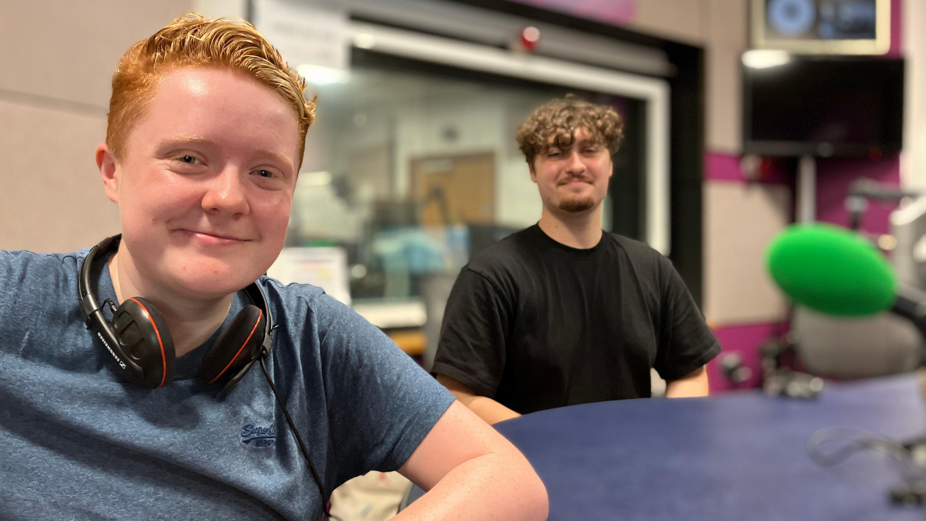 Two young people – one in the foreground and one in the background – sitting at a radio studio desk with headphones and a microphone. They are both smiling at the camera.