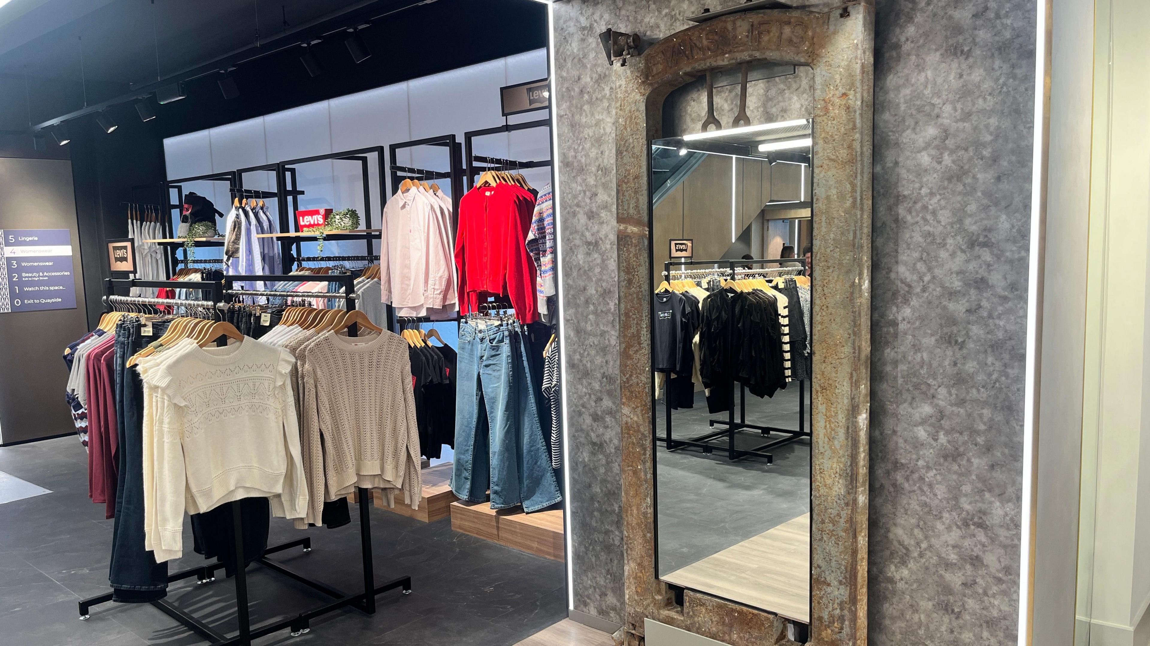 A mirror in the centre of a historic iron counterweight from a lift, on the wall of a modern clothing store.