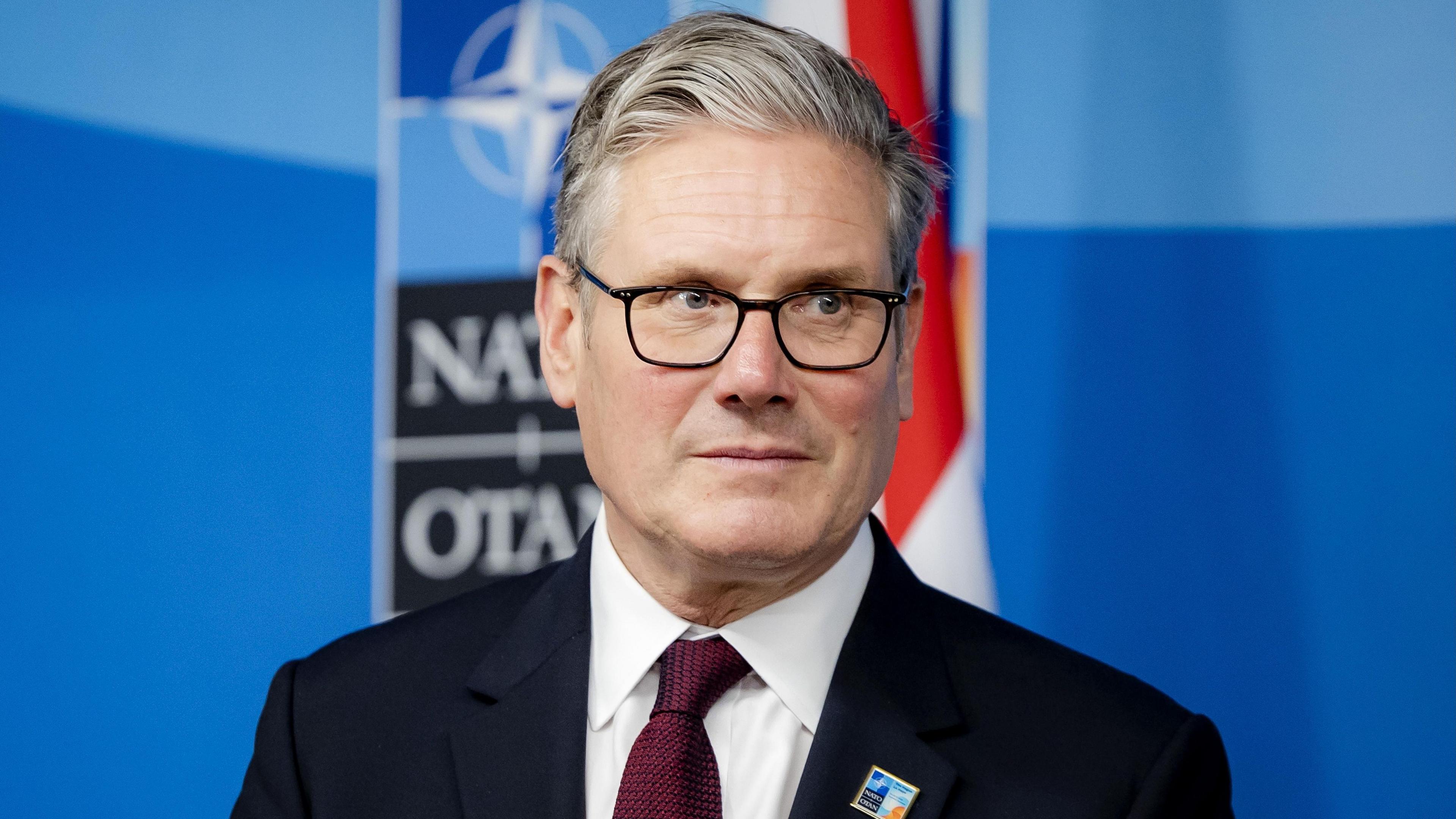 Prime Minister Sir Keir Starmer stands in front of a blue wall with the Nato emblem on it.