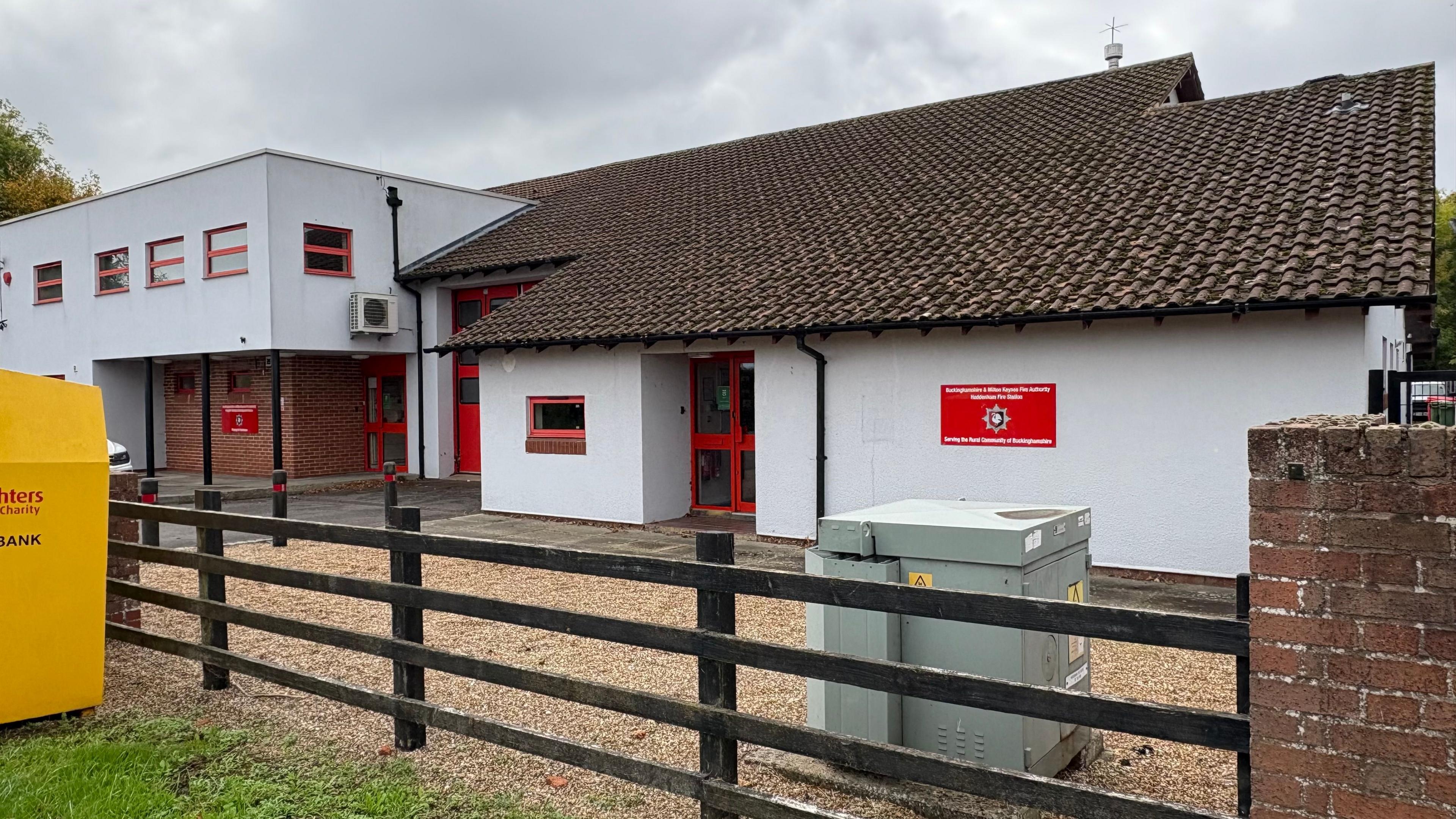 Single-storey white-painted building with red-framed window and doors to the right and two-storey white-painted building with red-framed windows and a flat roof to the left. There is a wooden fence near the camera and a yellow clothing bank skip to the left.