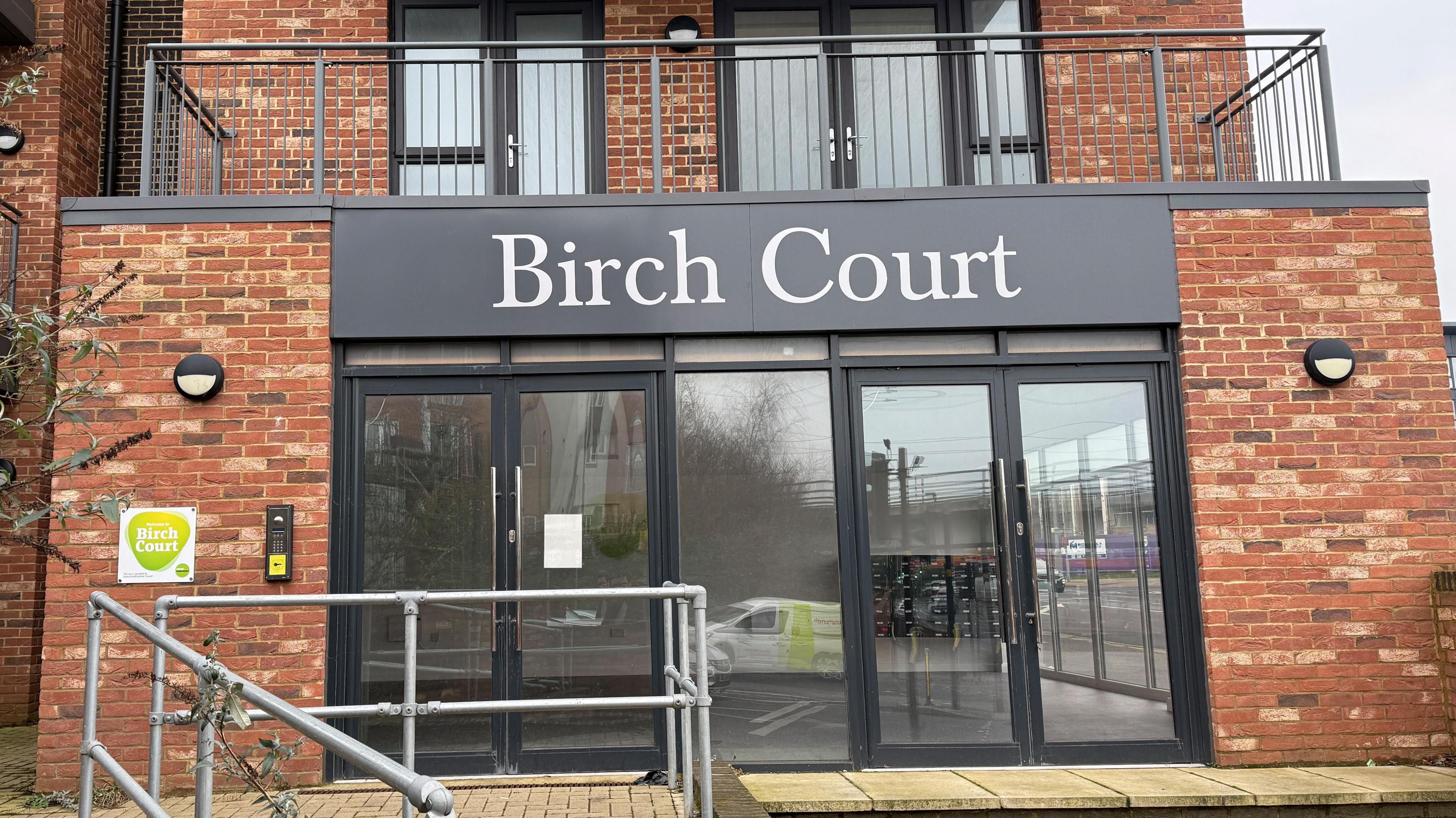 The main entrance to Birch Court is two sets of glass double-doors. There is an accessible ramp entrance on the left and steps on the right. The name "Birch Court" is displayed on a large grey sign in white font above.