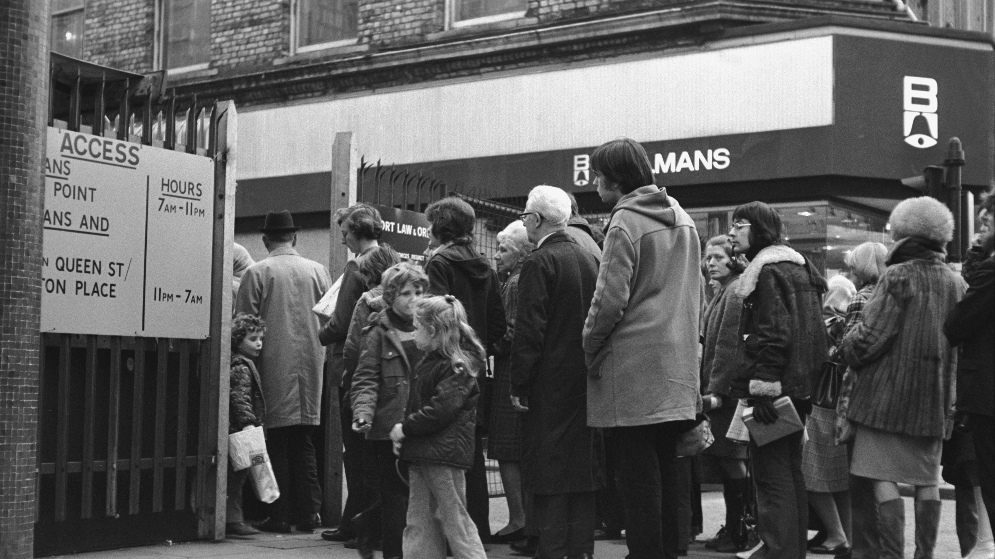 A black and white archive image of members of the public, including children, queuing outside a metal fence to enter Belfast city centre in 1972. A sign on the fence indicates the access point operates between 07:00 and 23:00. 