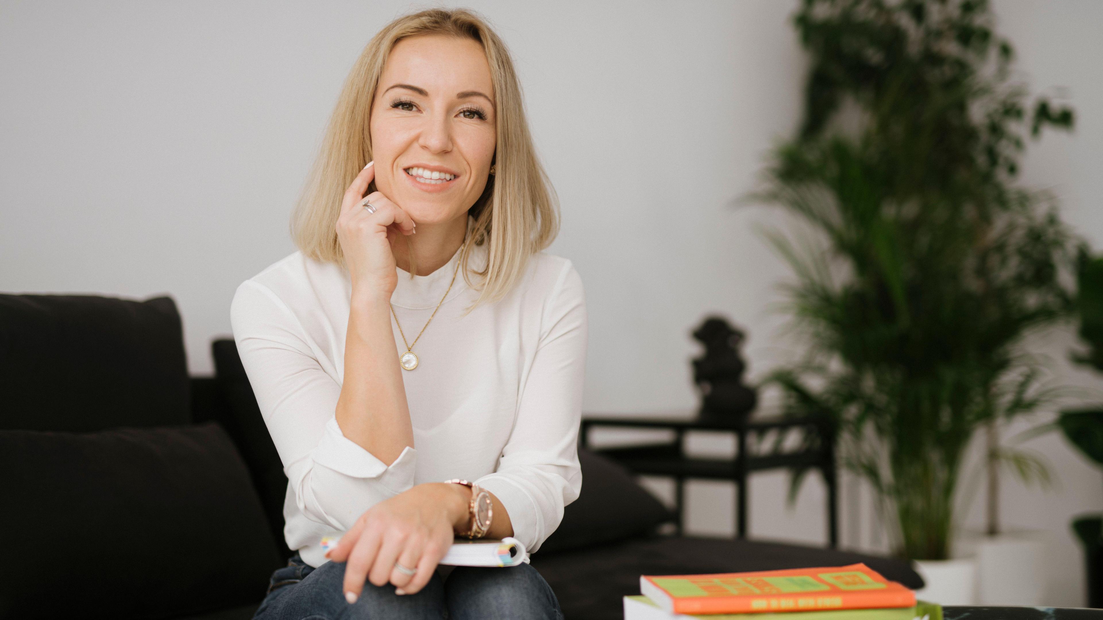 Aleksandra Pamplett sitting down with her arms folded across a book on her lap. She is blonde and wearing a white blouse and blue jeans