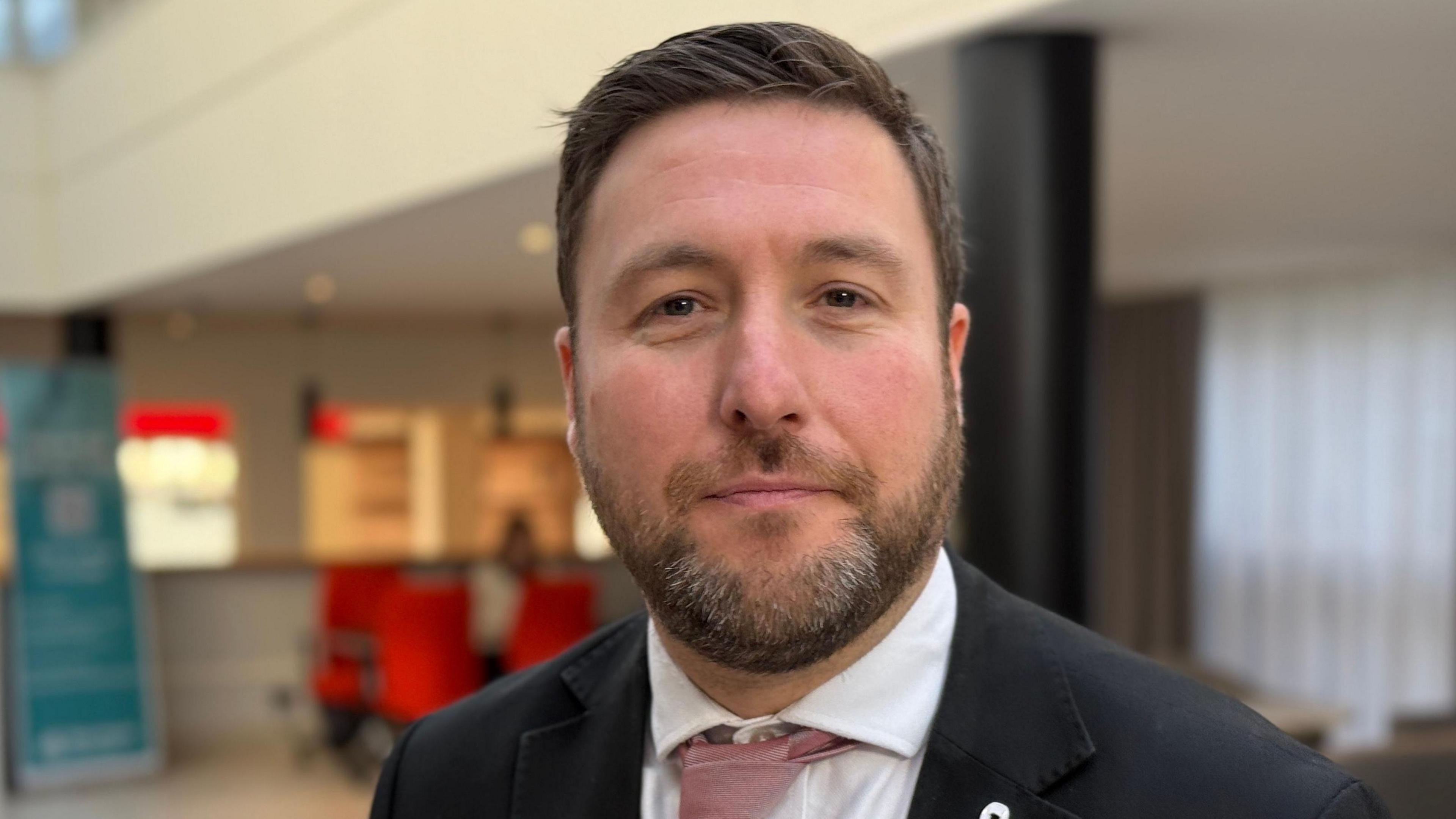 A picture of a man who has short dark brown hair and a dark brown beard with white bits. He is wearing a black suit jacket with a dark pink tie.