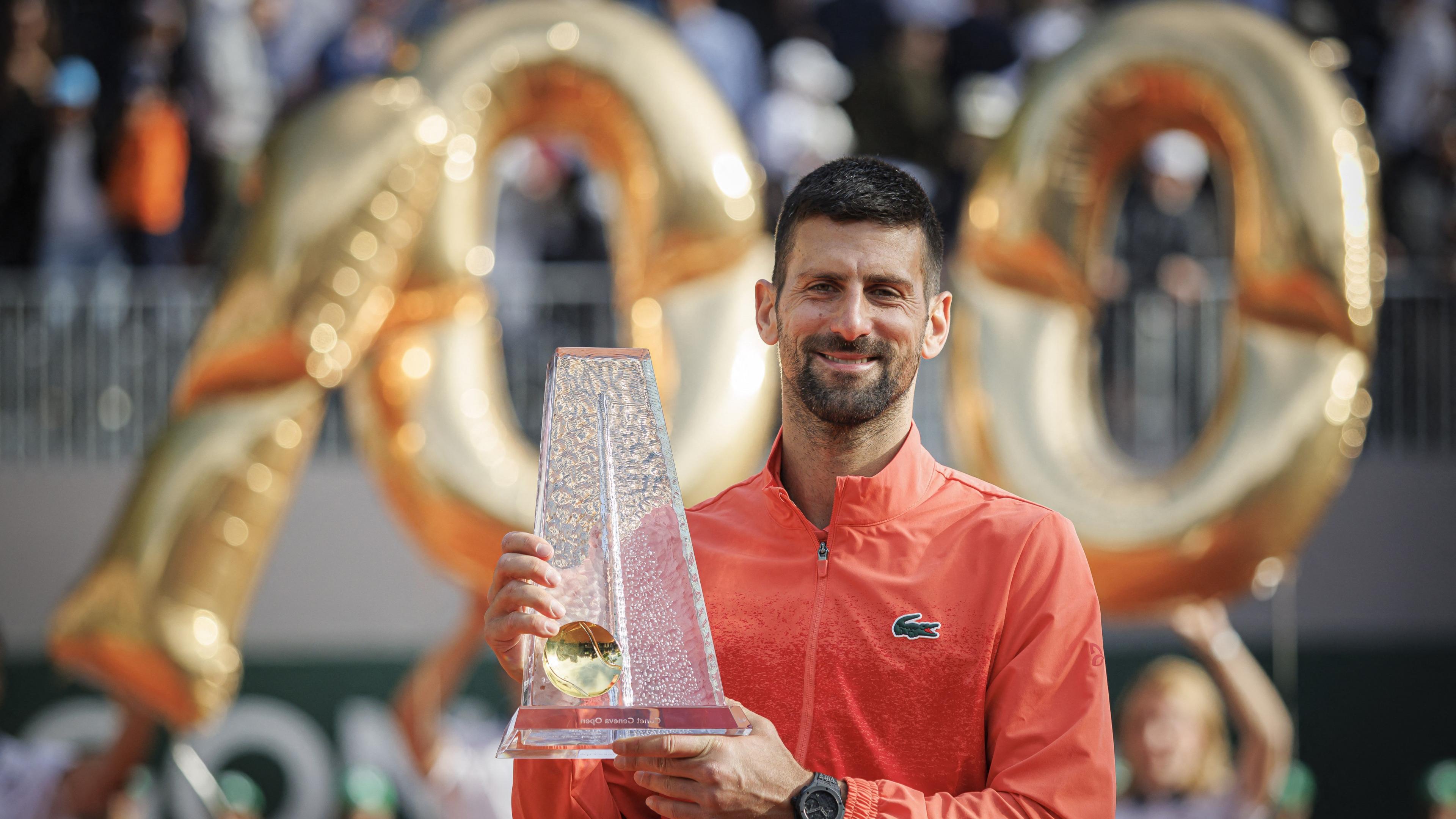 Novak Djokovic holds the Geneva Open trophy while balloons spelling out '100' hover in the background