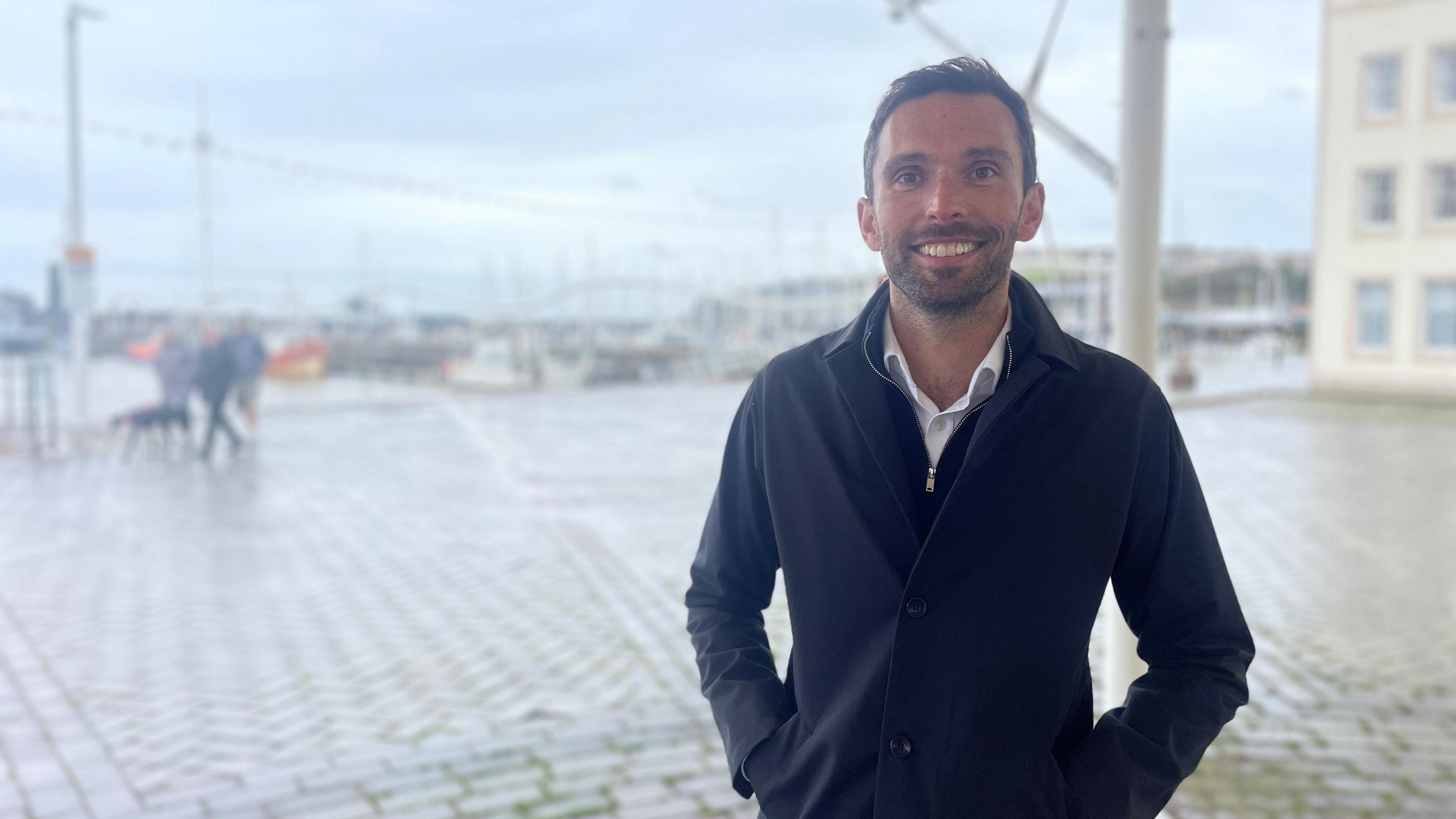 MP Josh MacAlister looking into camera and smiling under a gazebo on Whitehaven Harbour with a blurred background. 