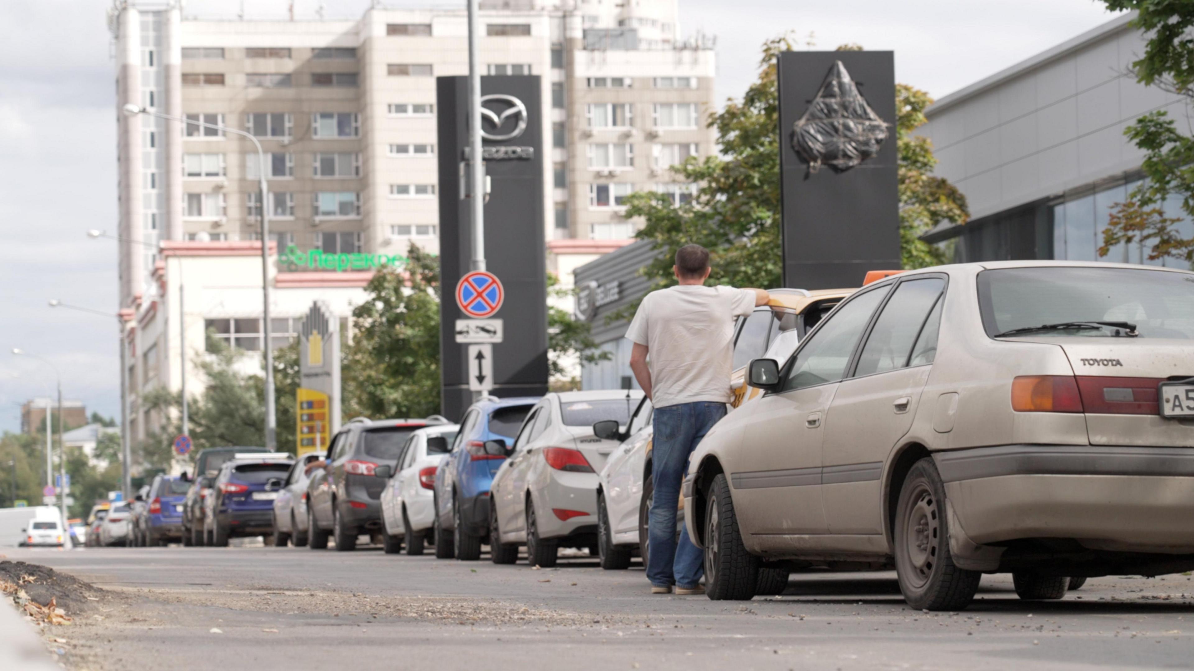 A man in a white t-shirt facing away from the camera stands with his right arm resting on his car, in a long queue of vehicles outside a petrol station in Moscow