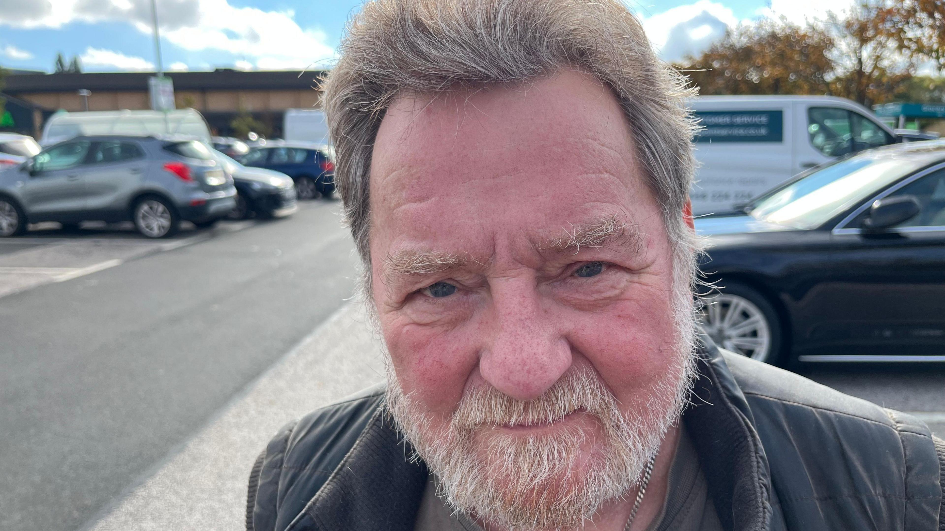 Peter Bone smiling at the camera in the same car park. He has grey hair and a beard and moustache. He is wearing a black puffer jacket.