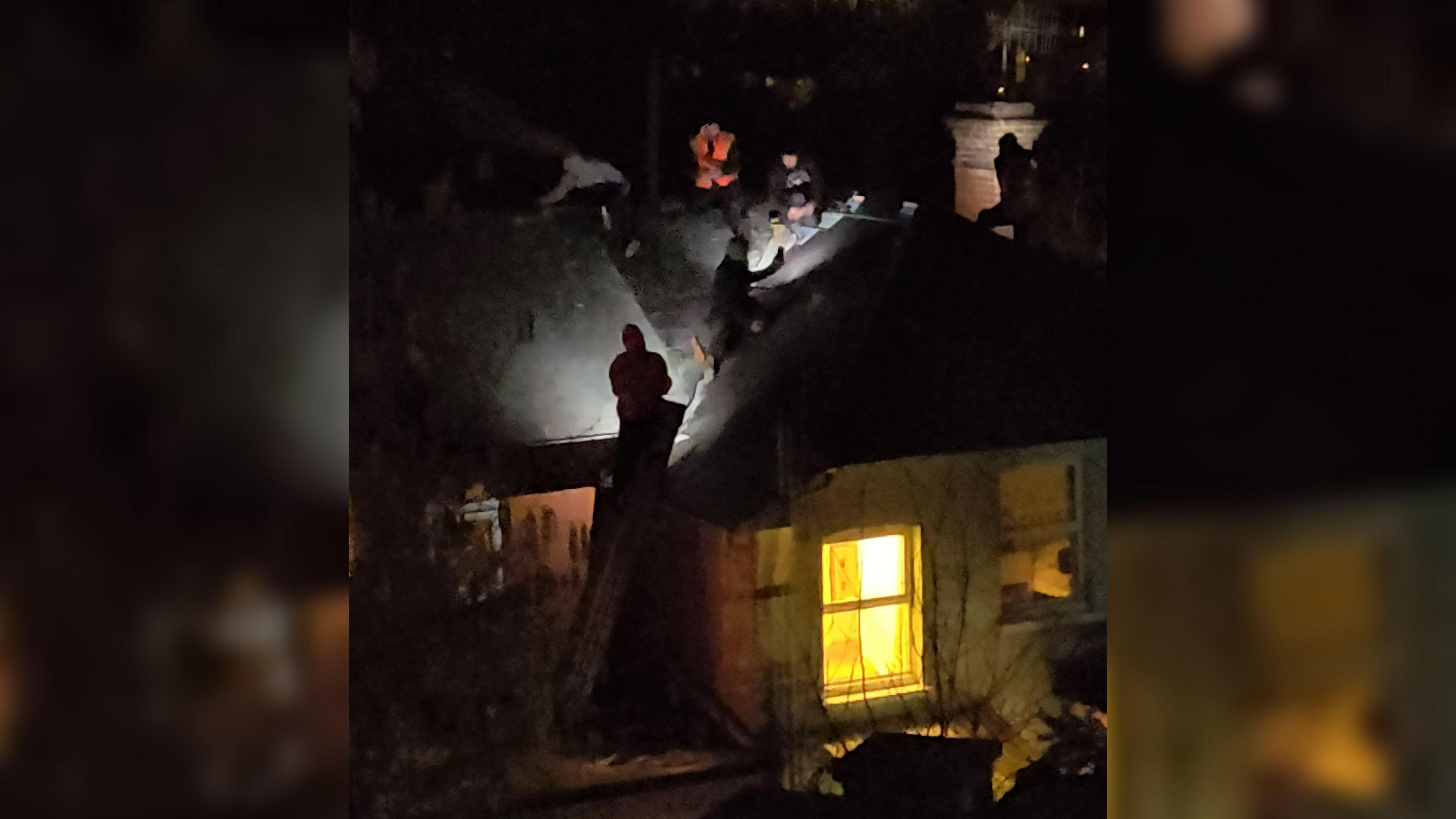 A night time picture showing a house in darkness and on the roof several people can be seen holding lights in their hands to illuminate different areas of the roof.