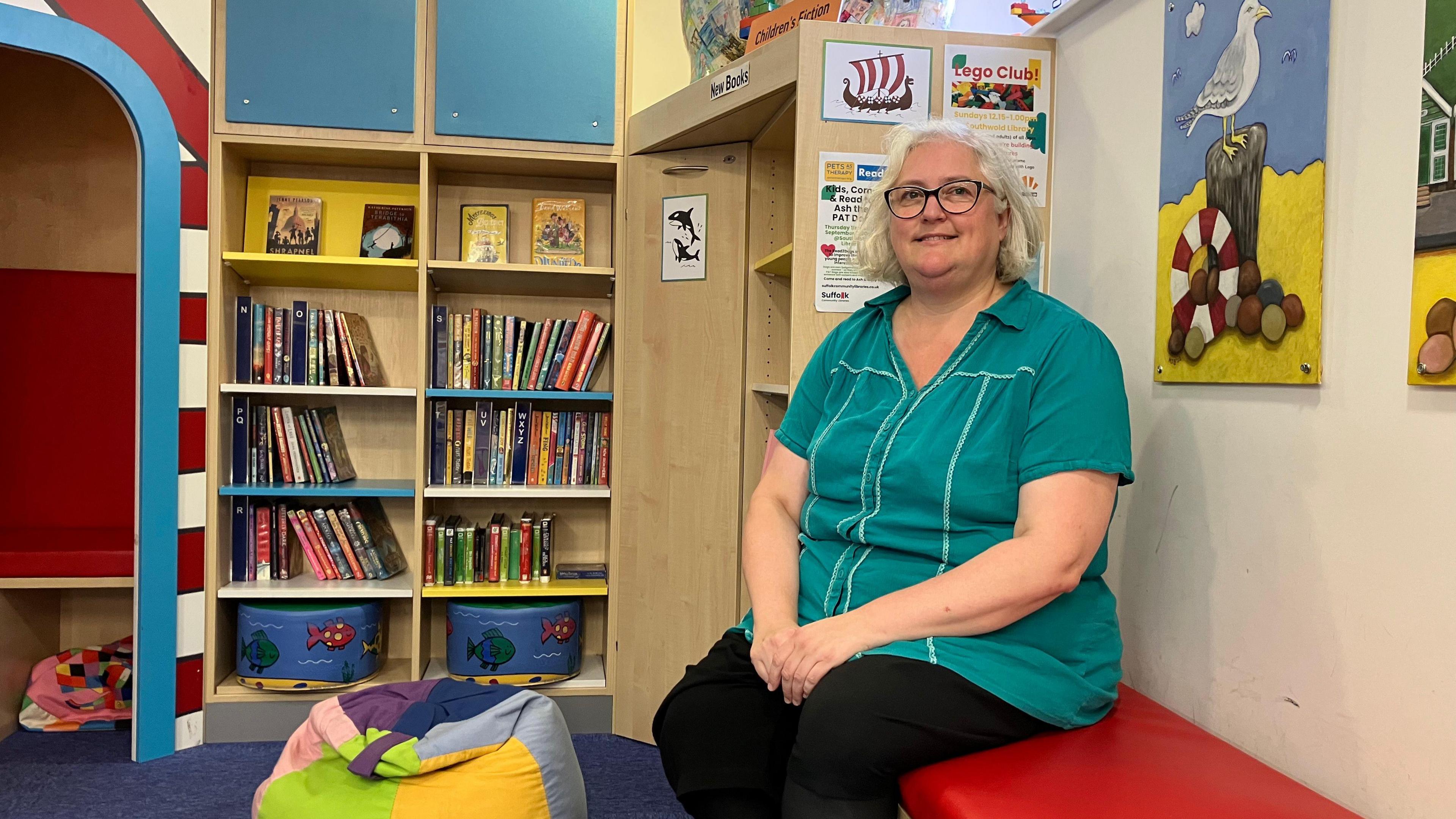 Charlotte Clark  sits on a bench with bookshelves and colourful children's posters on the wall.