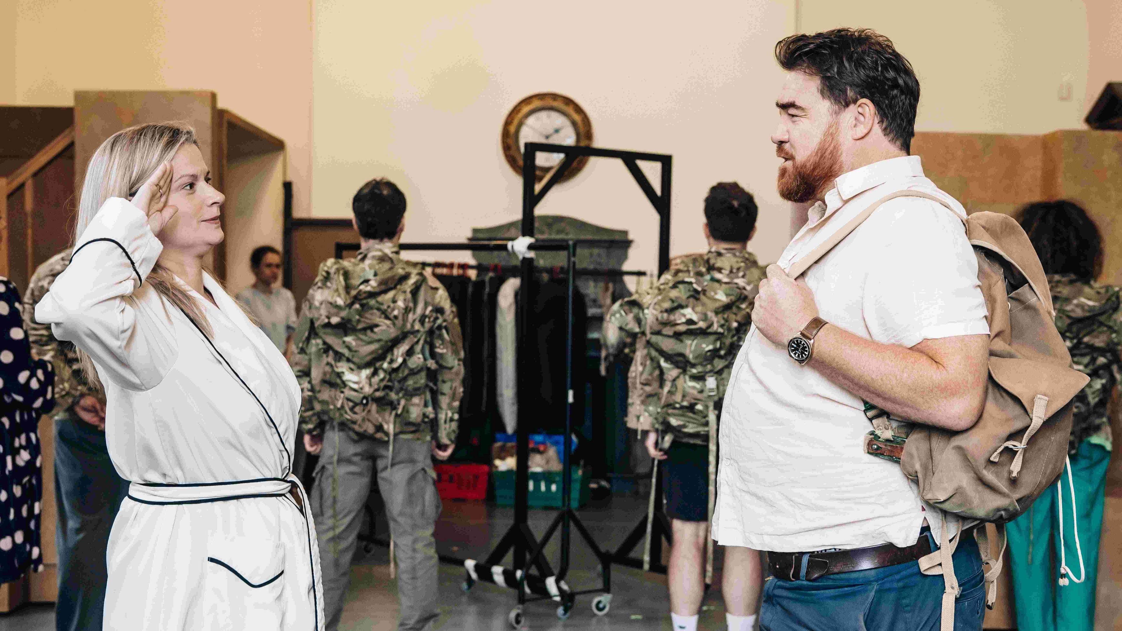 A rehearsal room for a theatre show. A woman salutes a man, playing the role of her husband. Soldiers are in the background, facing away from the camera.