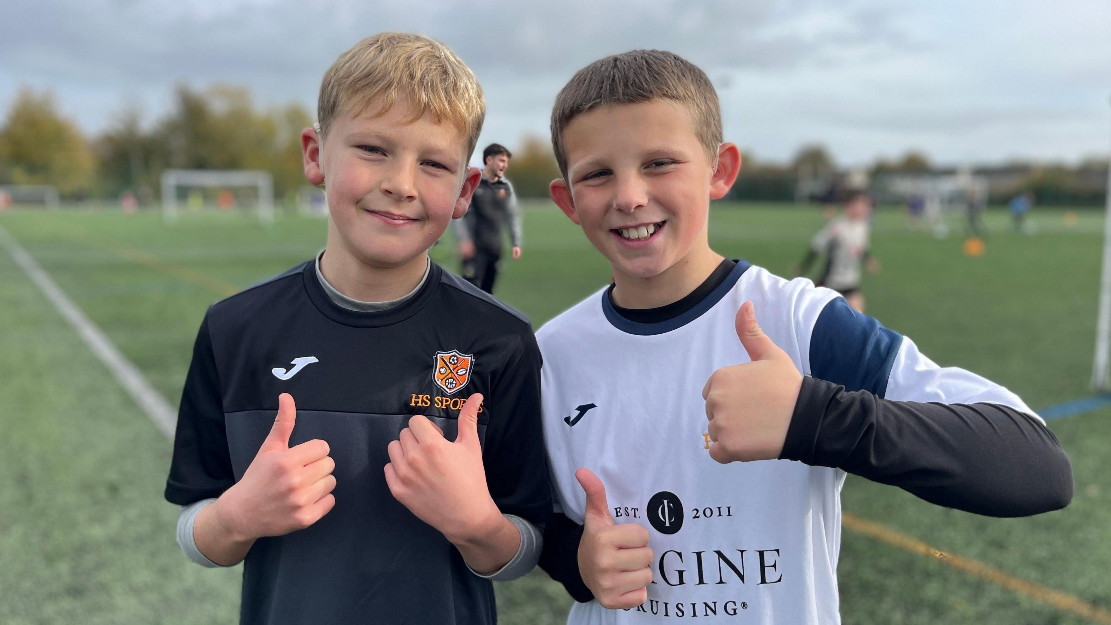 Stanley and Jacob stood next to each other on a football pitch. They are wearing football shirts and doing a thumbs up to the camera. They are both smiling and are stood by the corner flag on the football pitch. Behind them are other players on the pitch, but who are out of focus.