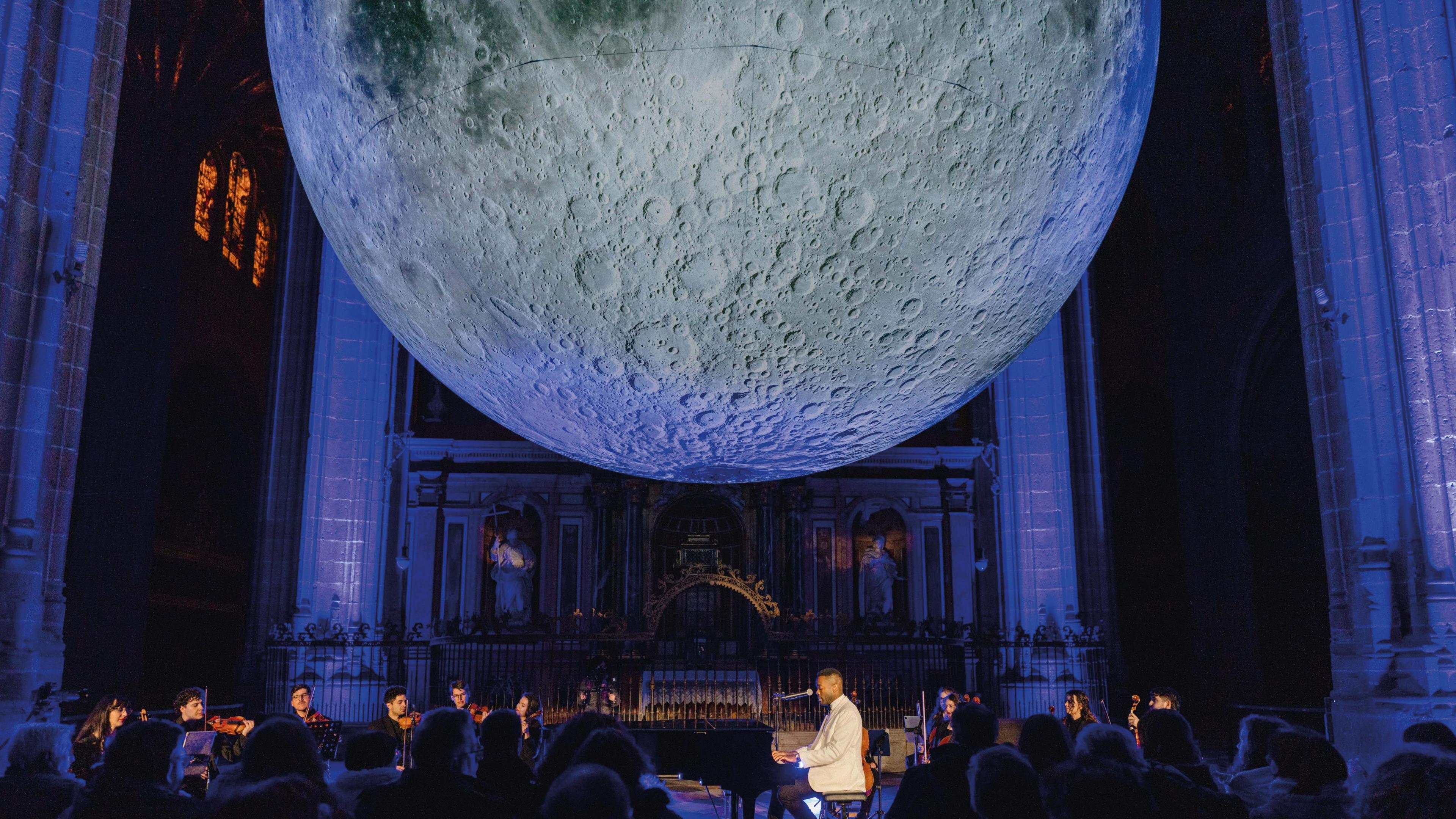An orchestra with a pianist wearing a white tuxedo top sitting at a piano, playing music underneath a giant Earth sculpture. They are in a church, with an organ behind the orchestra. The light is mostly blue, with the heads of the orchestra members illuminated.