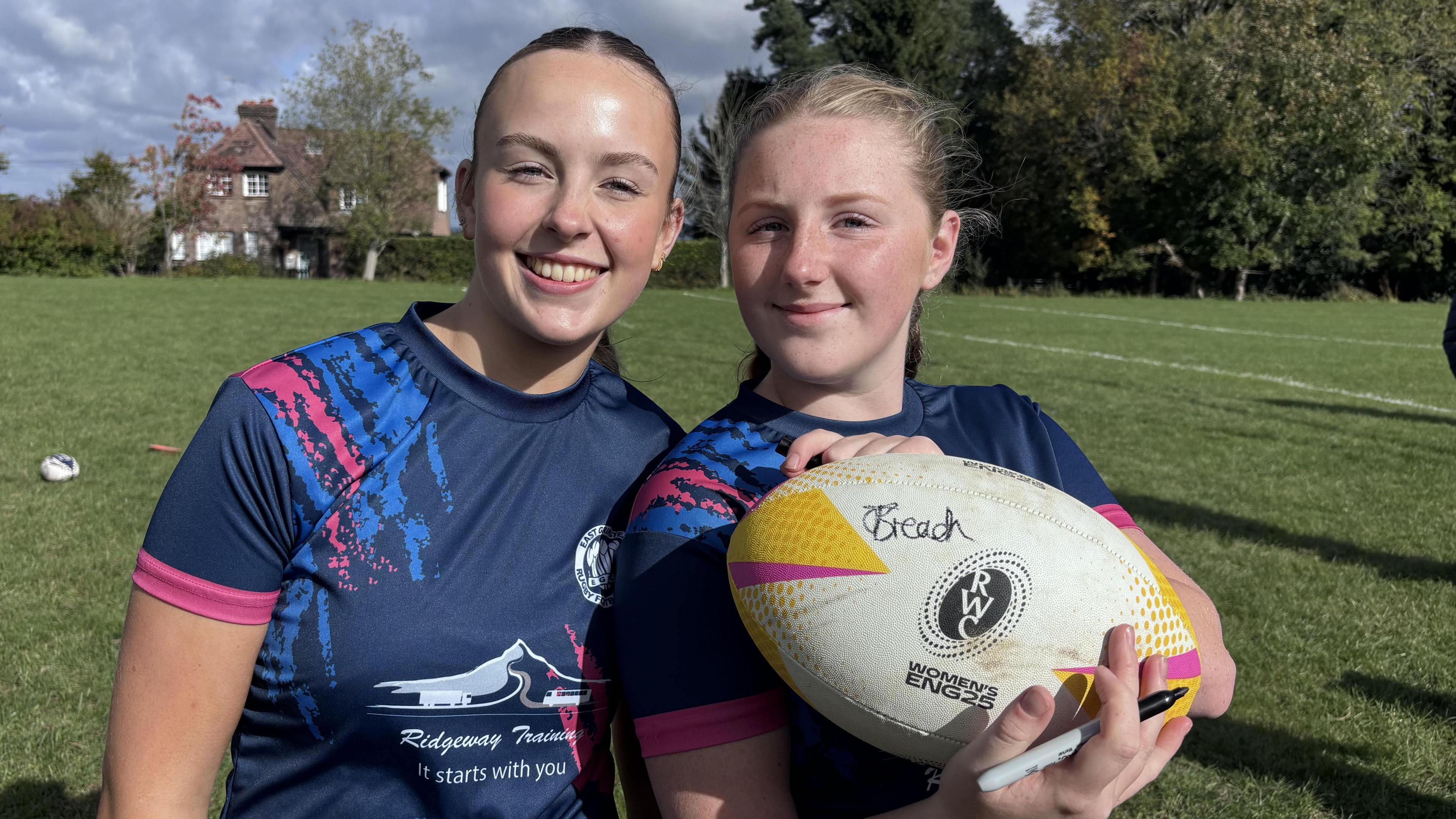 Millie and Izzie are wearing East Grinstead Rugby Club kits. Izzie is holding a rugby call which has been signed by Jess Breach.