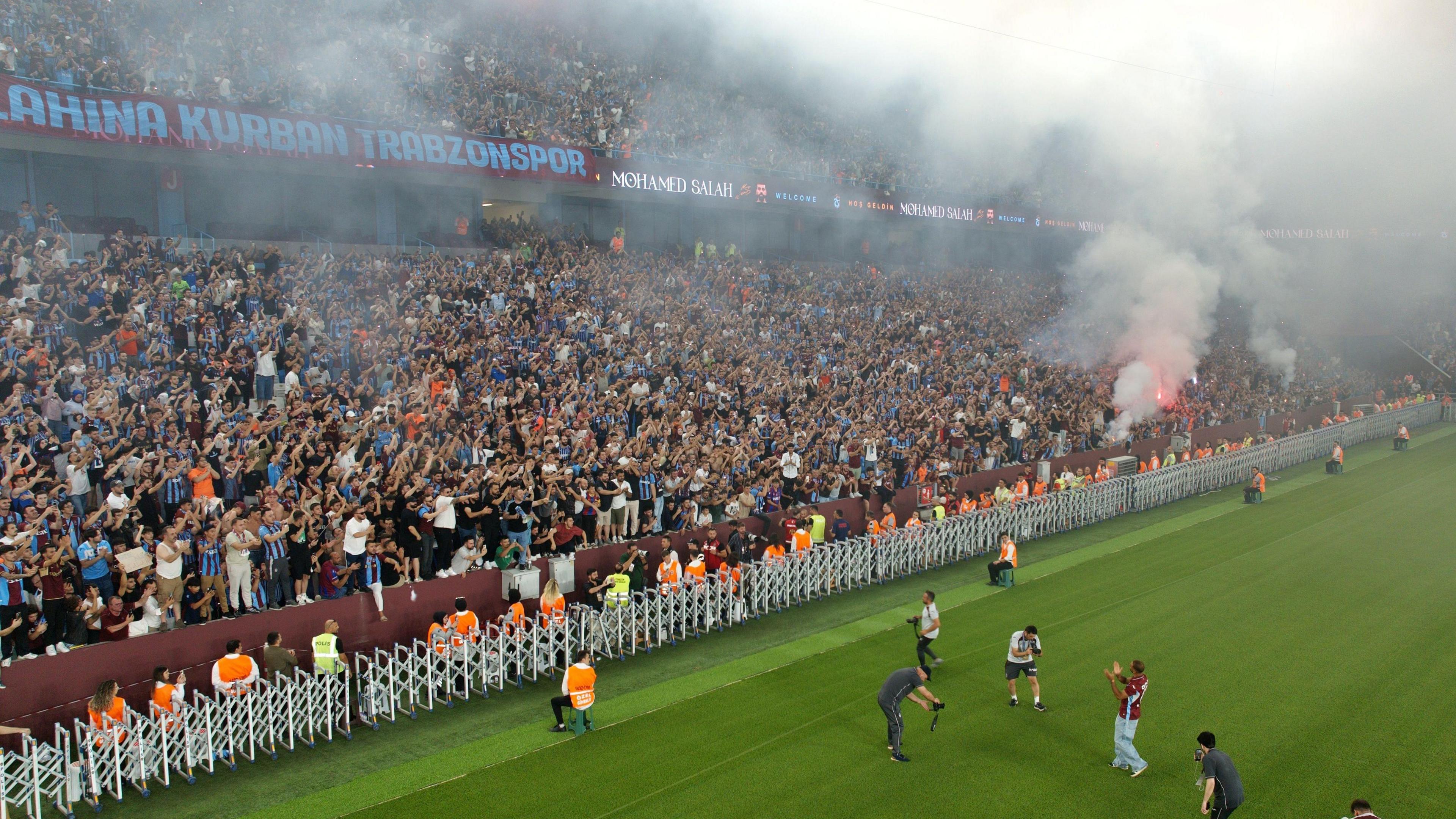 A drone shot shows a small-looking Mohamed Salah on the Papara Park pitch applauding in front of a vast stand filled with Trabzonspor fans, with smoke seen drifting from red flares. Several men with cameras gather around Salah to film and take pictures