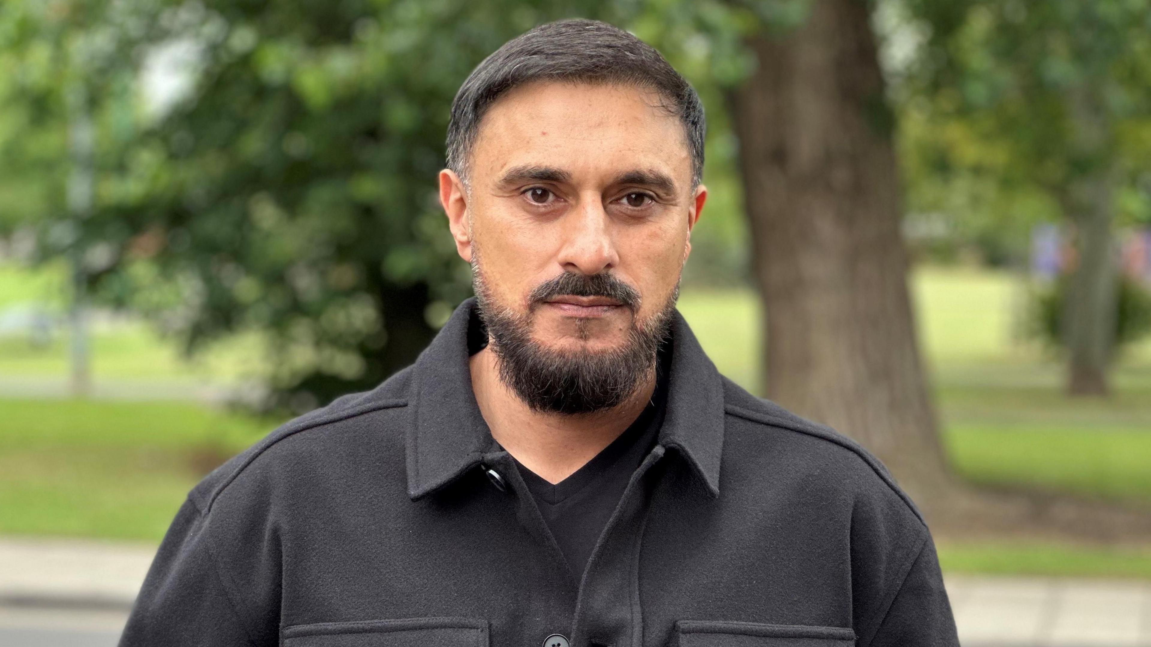 Amjid Khazir is stood in a black jacket near a park in Middlesbrough