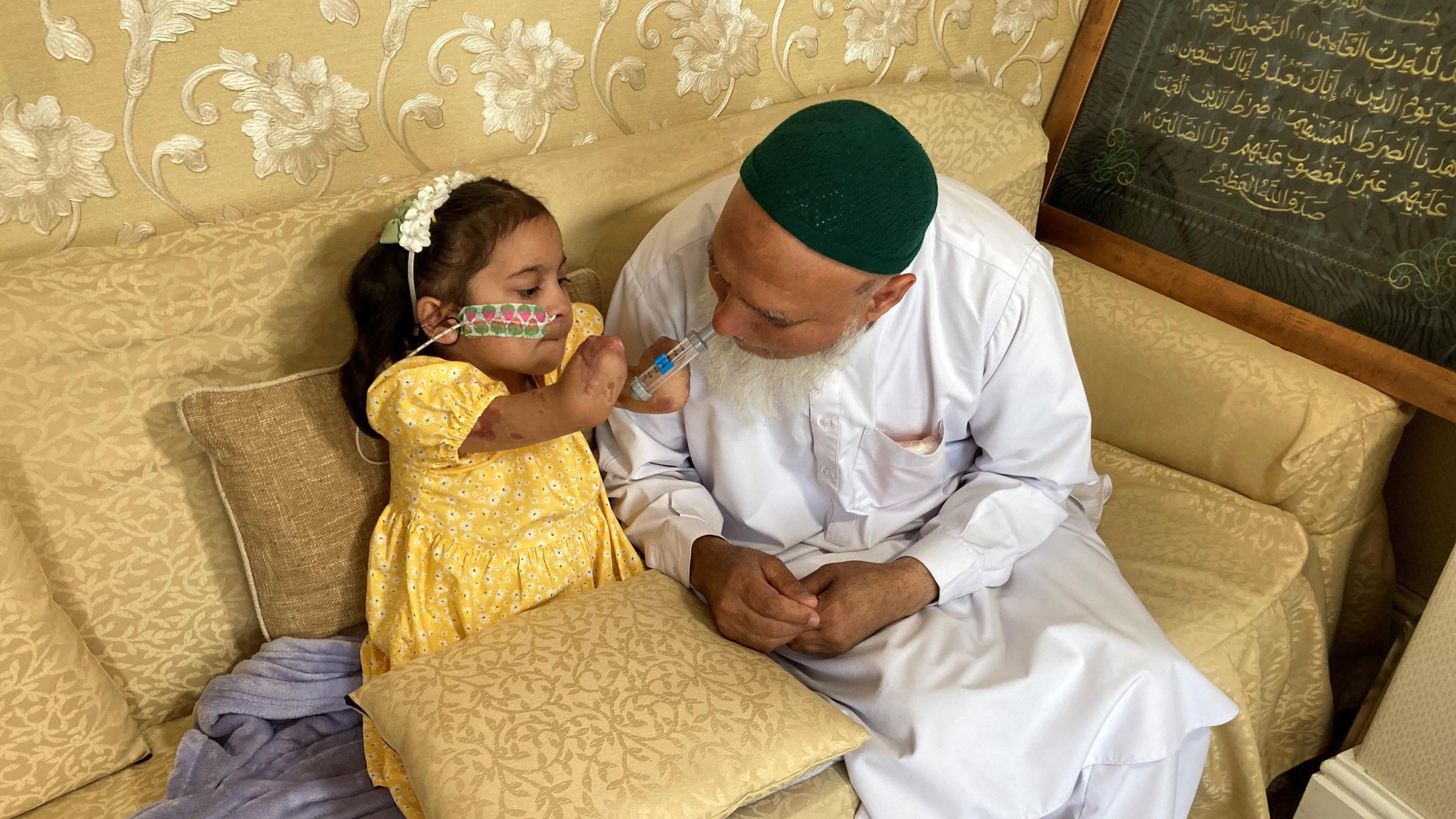 Saffanah sitting on a beige sofa with her grandfather. She is holding a toy syringe to his mouth and he is leaning down towards her.