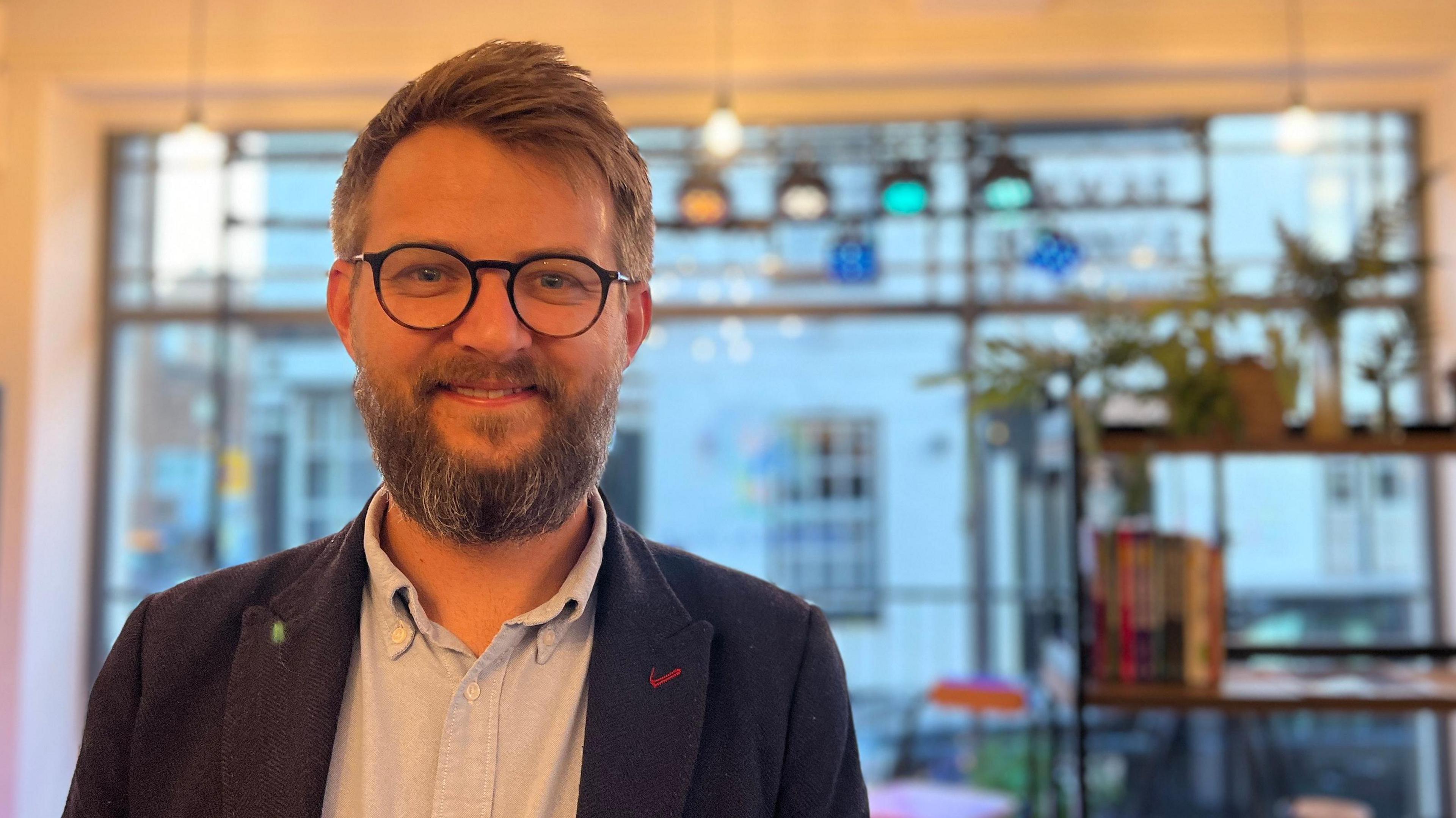 Alex Hughes is standing inside the community hub. He has a beard and circle-framed glasses and is wearing a navy blazer and a light blue shirt. He is smiling at the camera