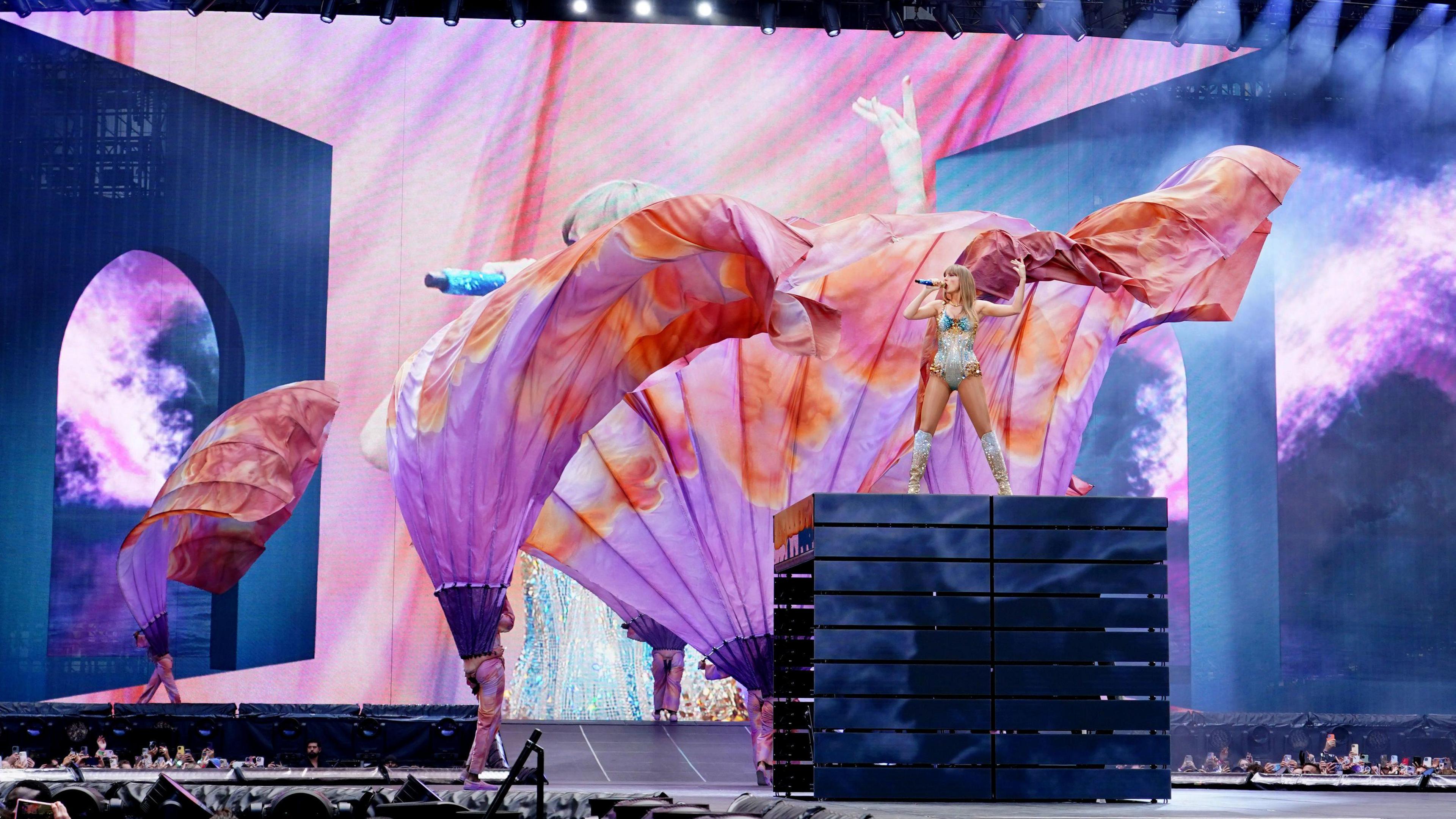 Taylor Swift performs on a platform high above Wembley stadium. Dancers are perfoming around her carrying large fabric banners.