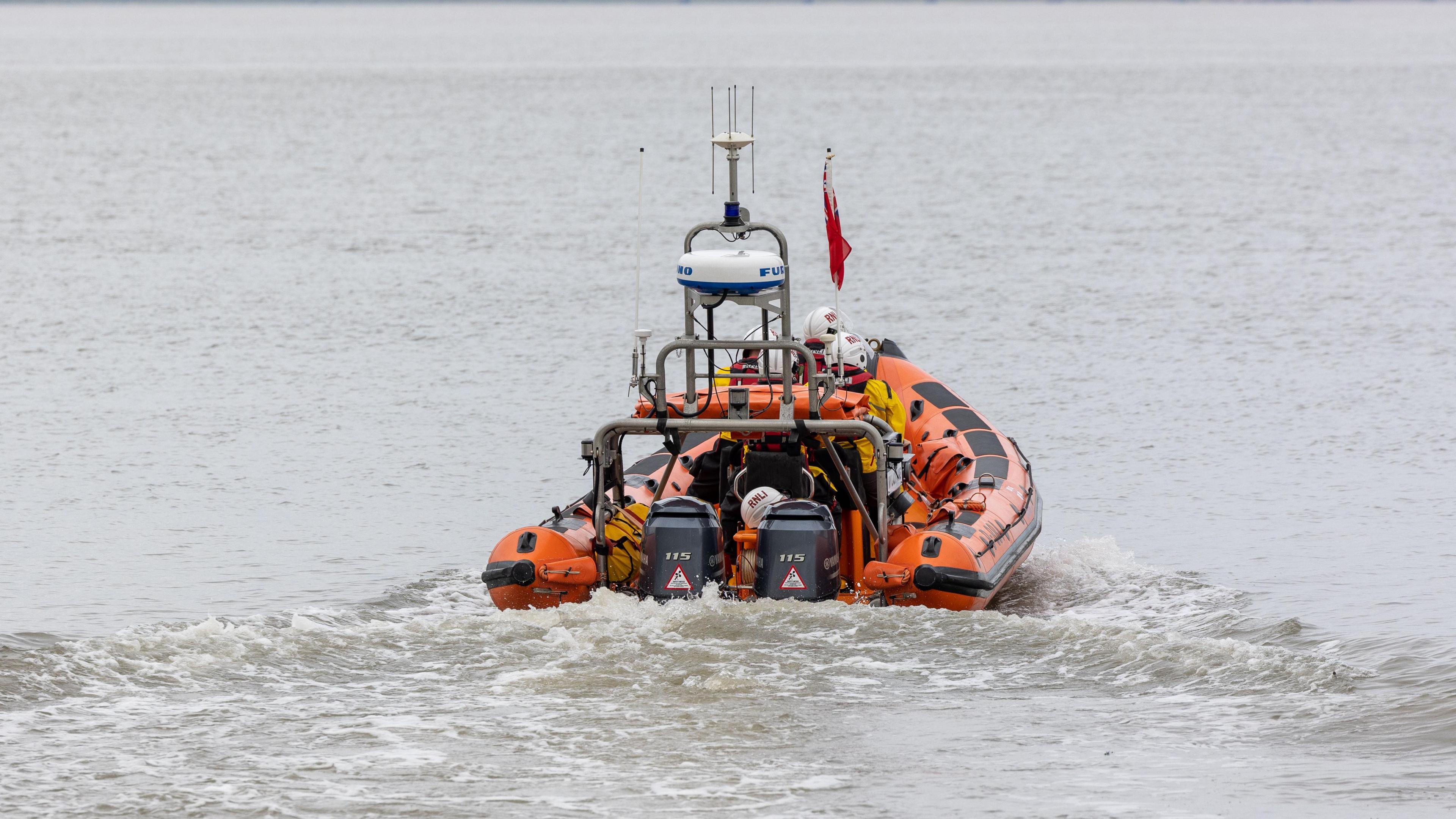 An orange RNLI boat at sea. A few crew members are onboard and there is a red flag on the front of the boat. They are travelling away from the camera, looking straight ahead. 