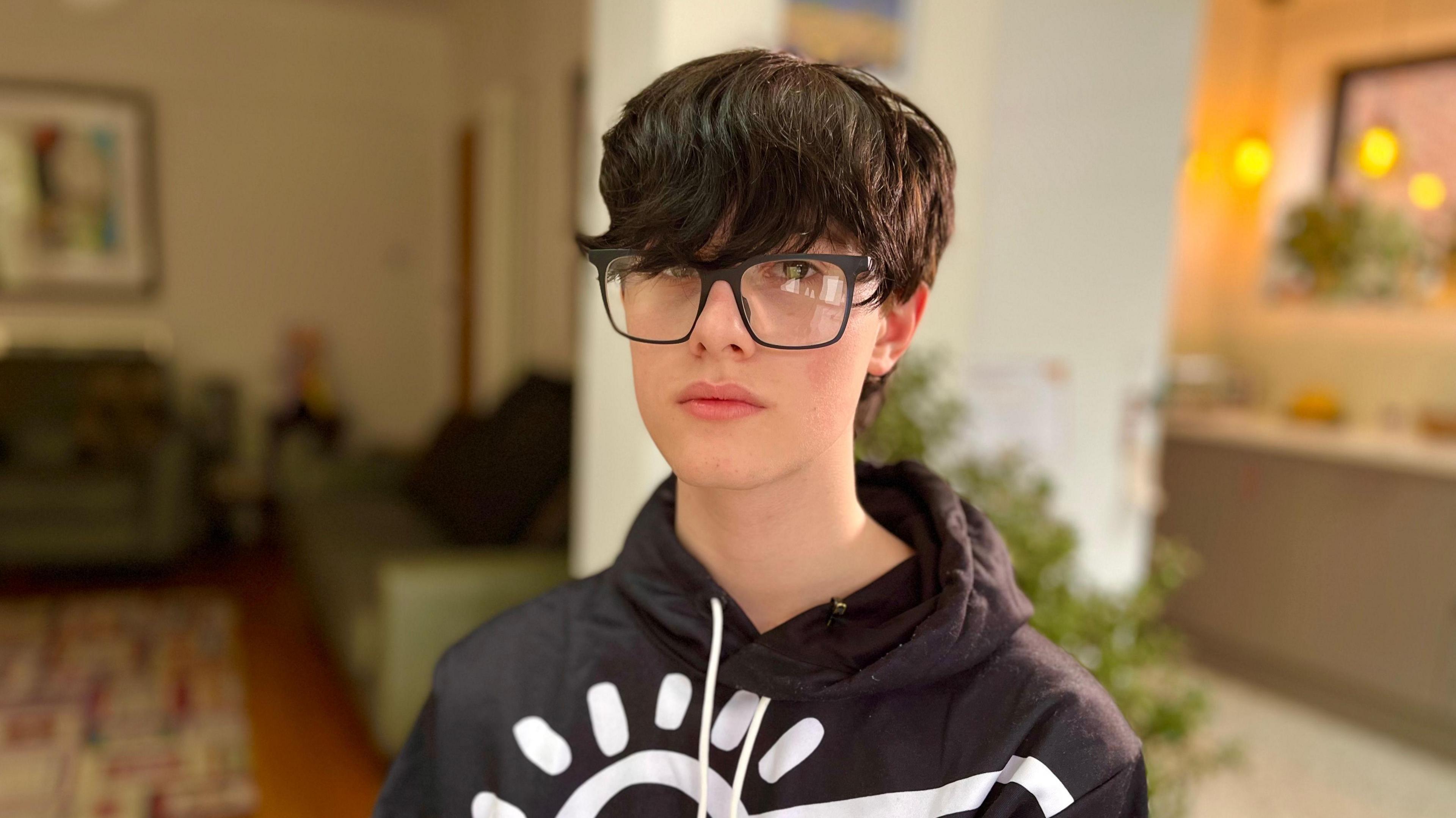 Head and shoulders photo of Michael wearing black rimmed spectacles and a black and white patterned top, sitting in a living room 