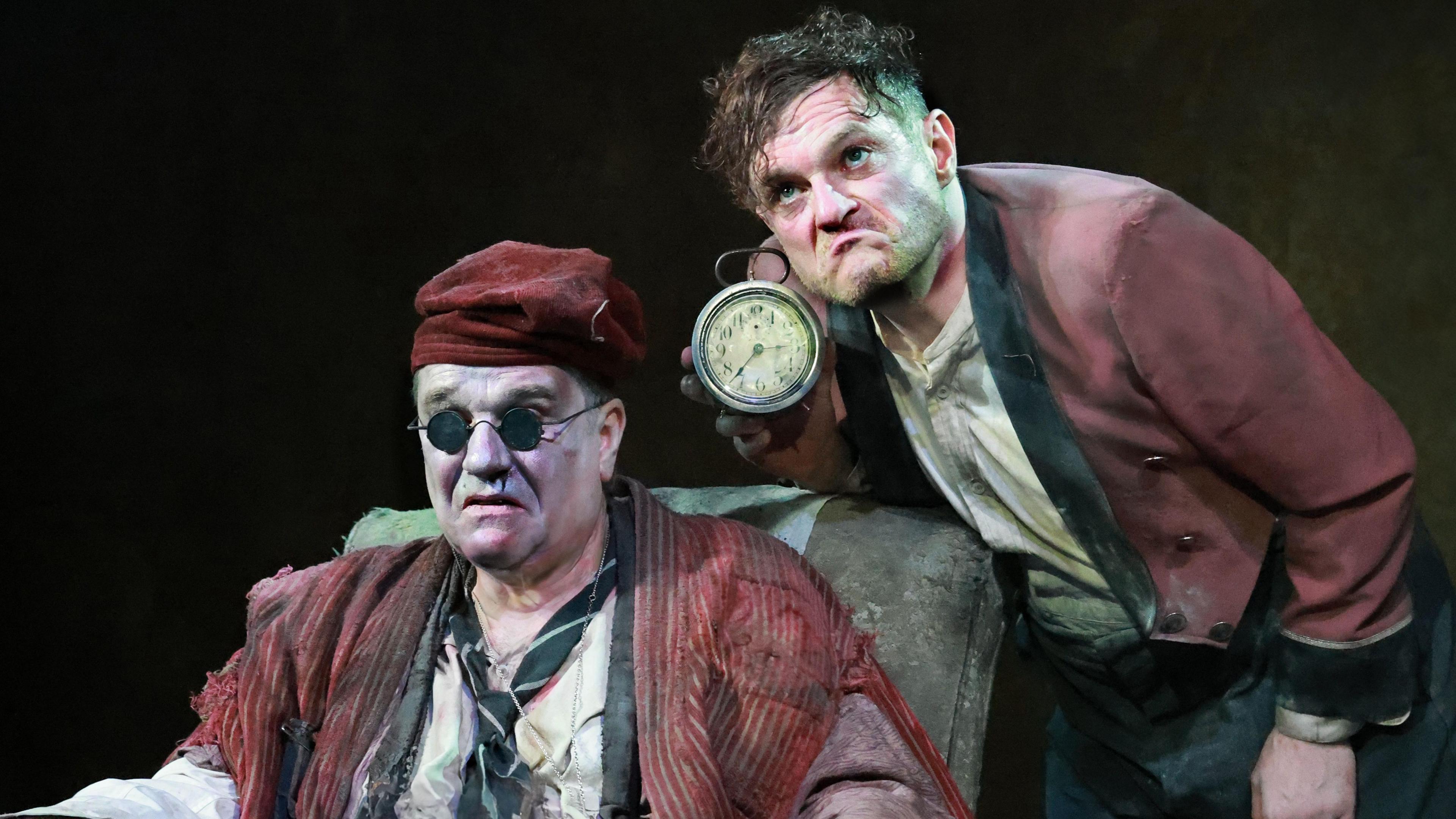 Two male actors on stage. One is sat on a chair wearing dark circular sunglasses and is wearing a red hat, a shirt and a tie. He is frowning. The other actor is behind him holding a clock. He is also frowning.