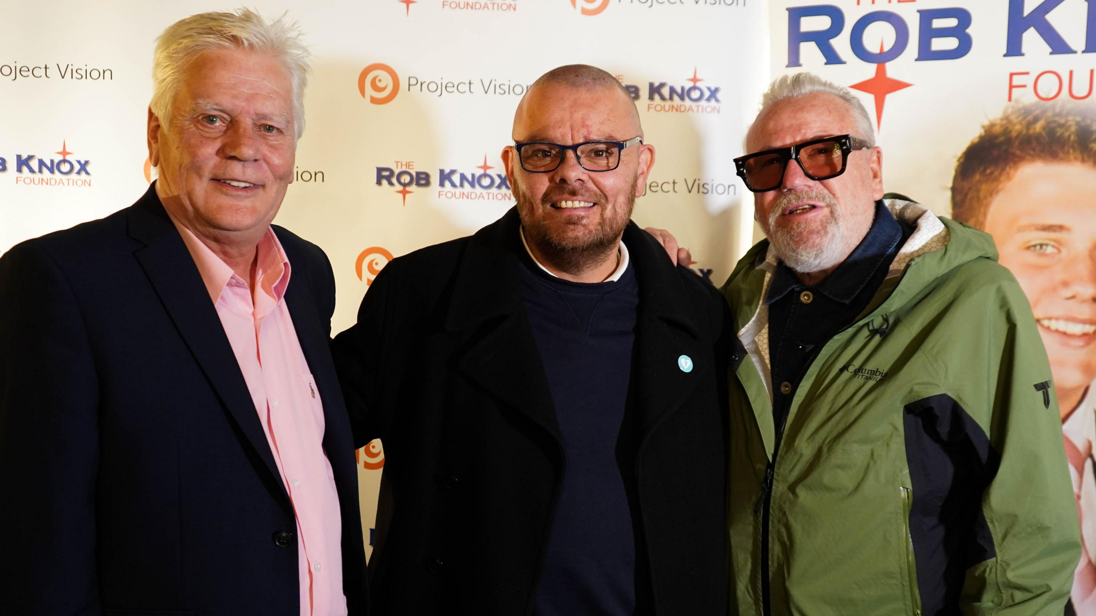 On the far left Mr Knox is a white-haired man in a black suit jacket and pink shirt. In the middle Mr Cosser has a shaved head and glasses is wearing a black  jacket and jersey - he has his arms around Mr Knox and Mr Winstone who is wearing a green jacket and tinted glasses. Behind them there is a step and repeat board which is branded with "the Rob Knox Foundation".