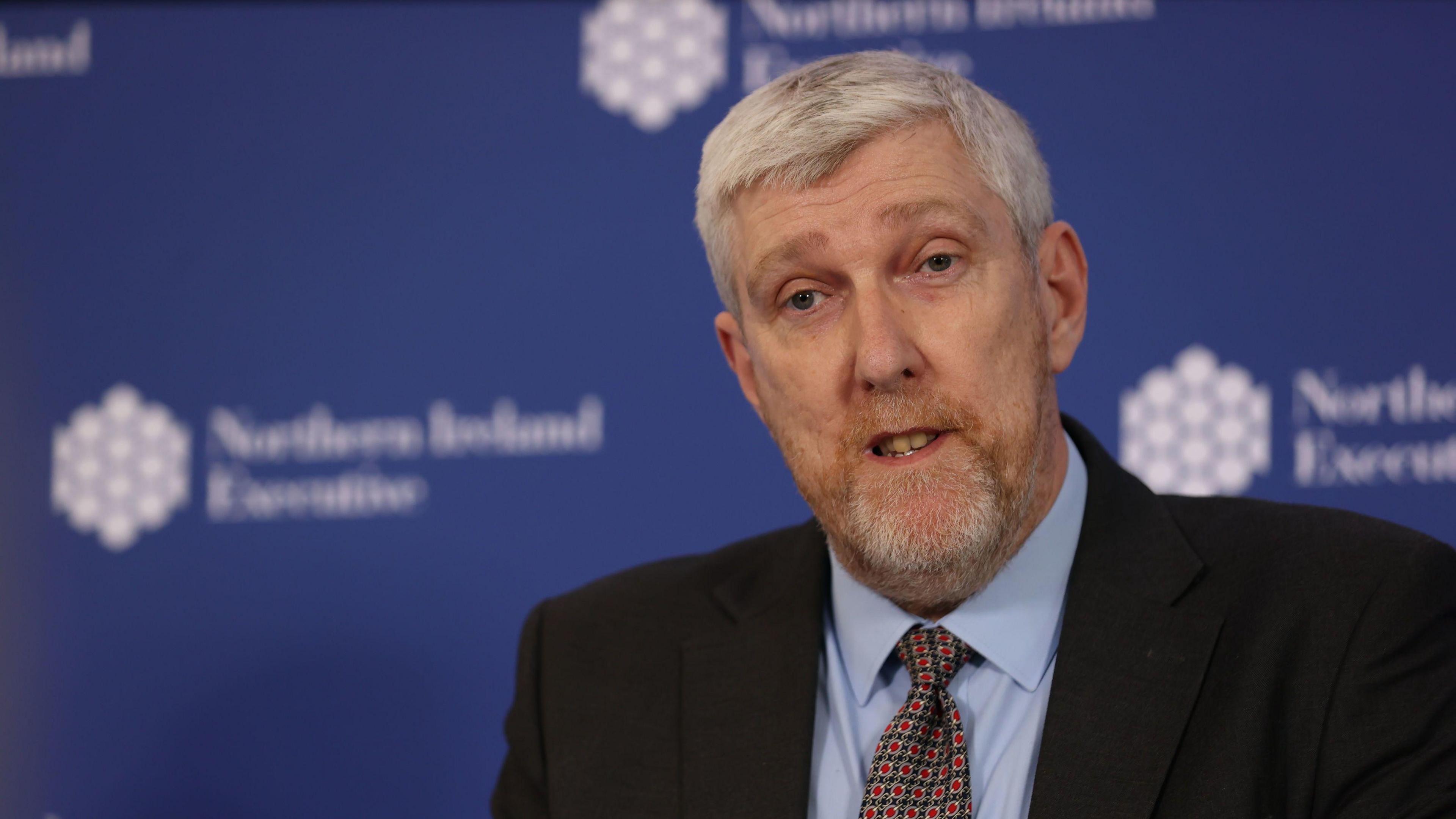 A man with grey hair and beard is wearing a black suit, blue shirt and a green and red pattern tie. He is in the bottom right of the frame and looks to be speaking. Behind him is a blue blurred background with the Northern Ireland Executive logo placed throughout.