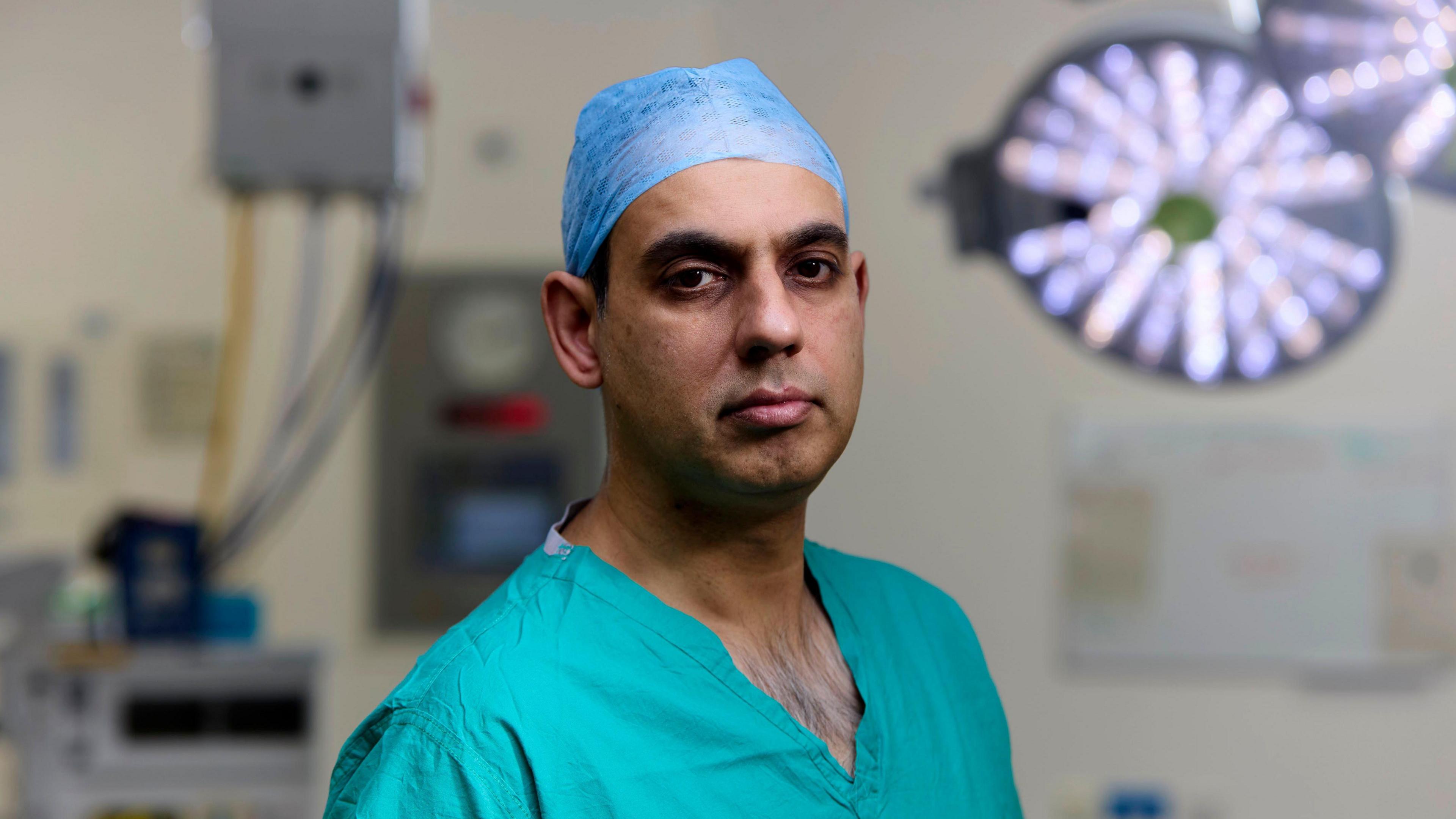 Surgeon Imran Liaquat adopts a serious look standing in the operating theatre, wearing turquoise blue scrums and theatre cap, the circular bright light behind him and monitors in the background