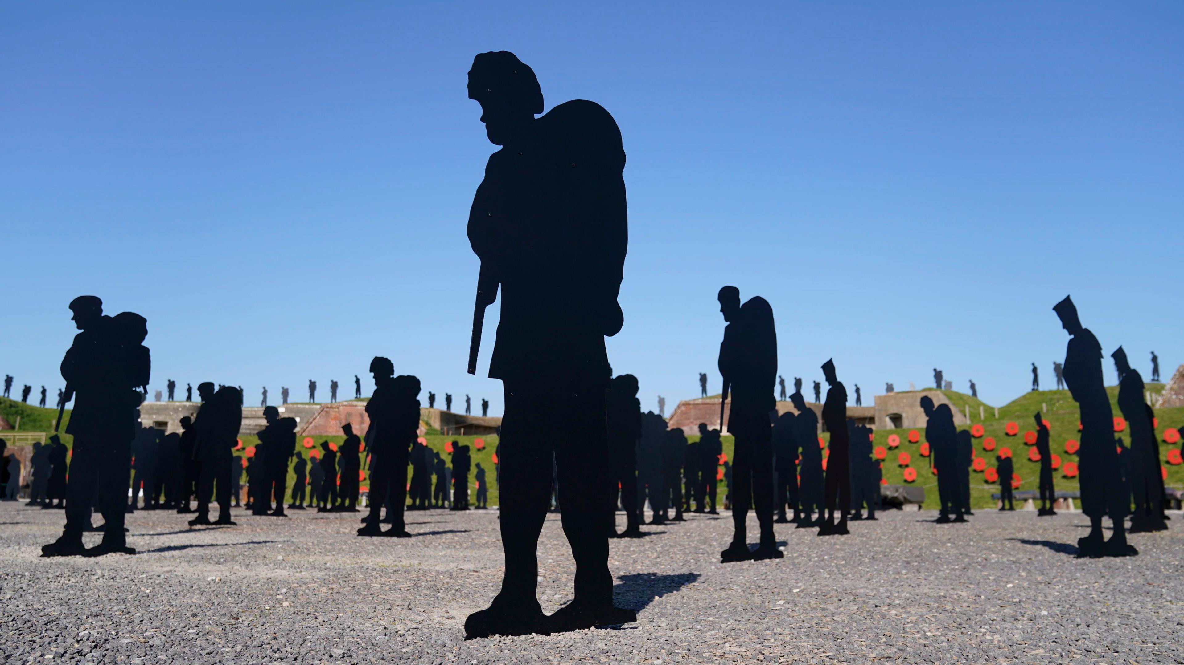 Hundreds of black silhouettes of soldiers in a gravel parade ground in the centre of a historical fort surrounded by grass banks covered in giant poppies