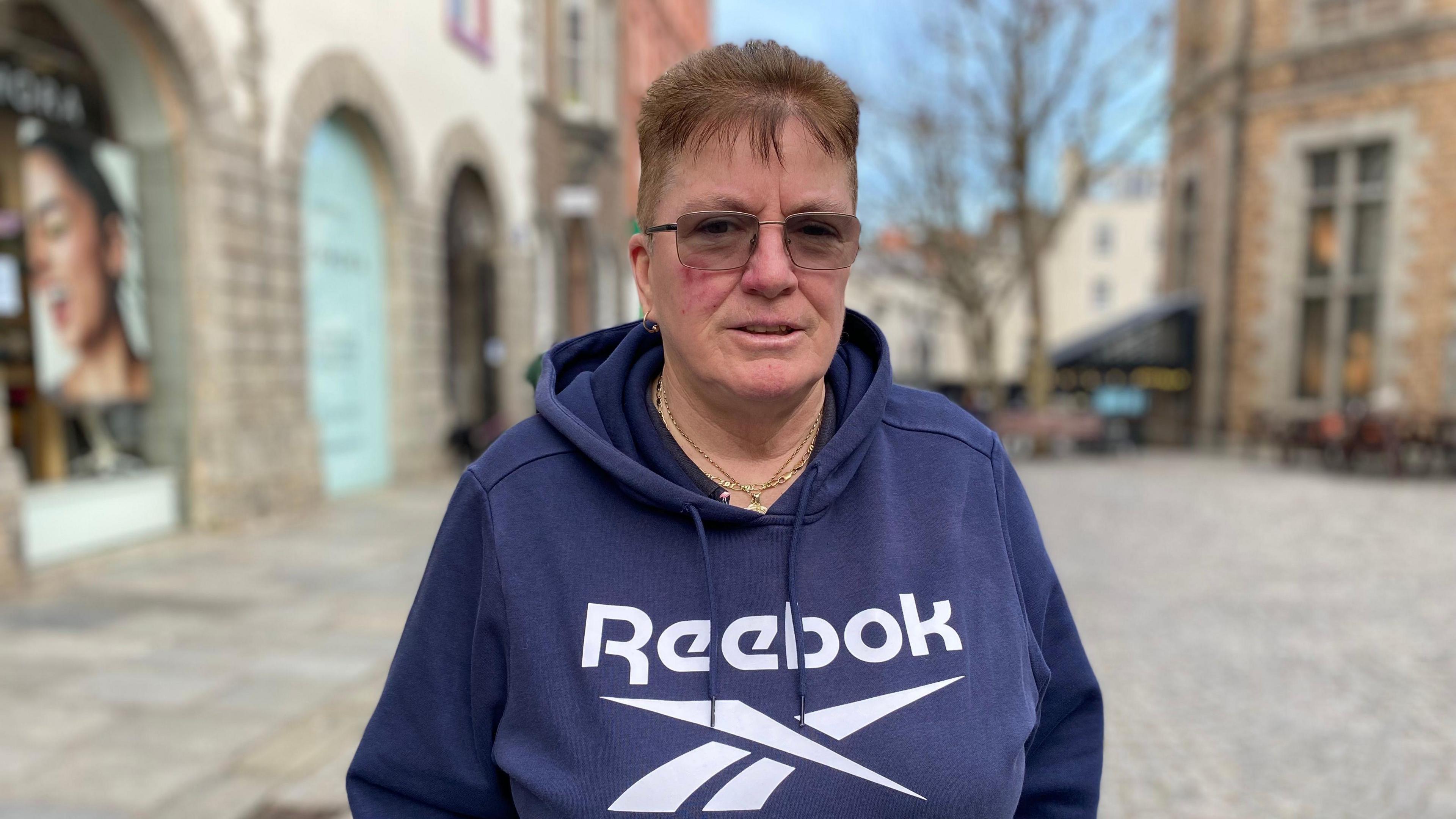 A woman with glasses and short brown hair in a blue hoodie jumper stands in Guernsey's Market Place.