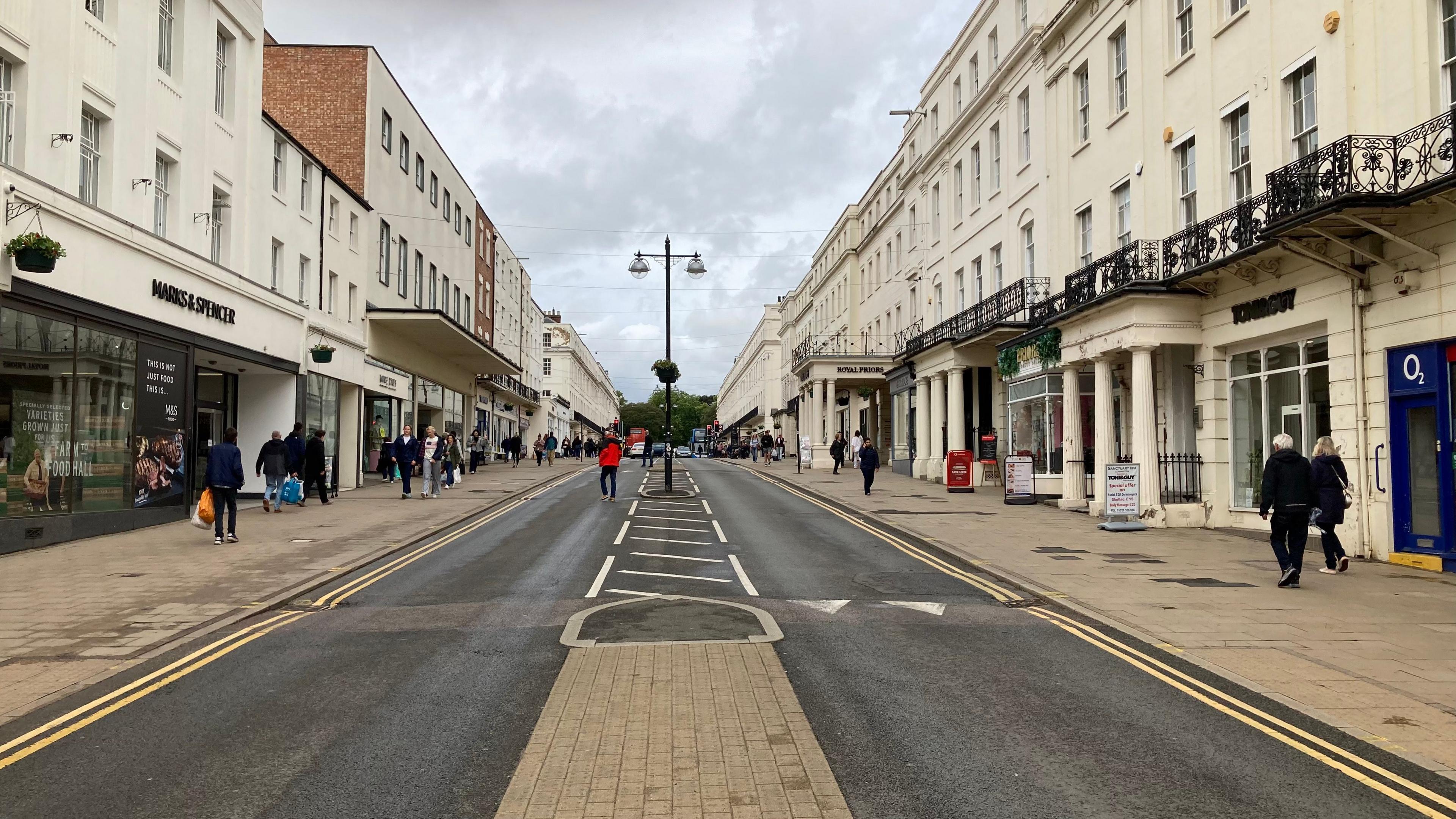 Leamington Spa High street with shops on either side of a long straight road. People can be seen walking on either side with one man in a red coat crossing the road.