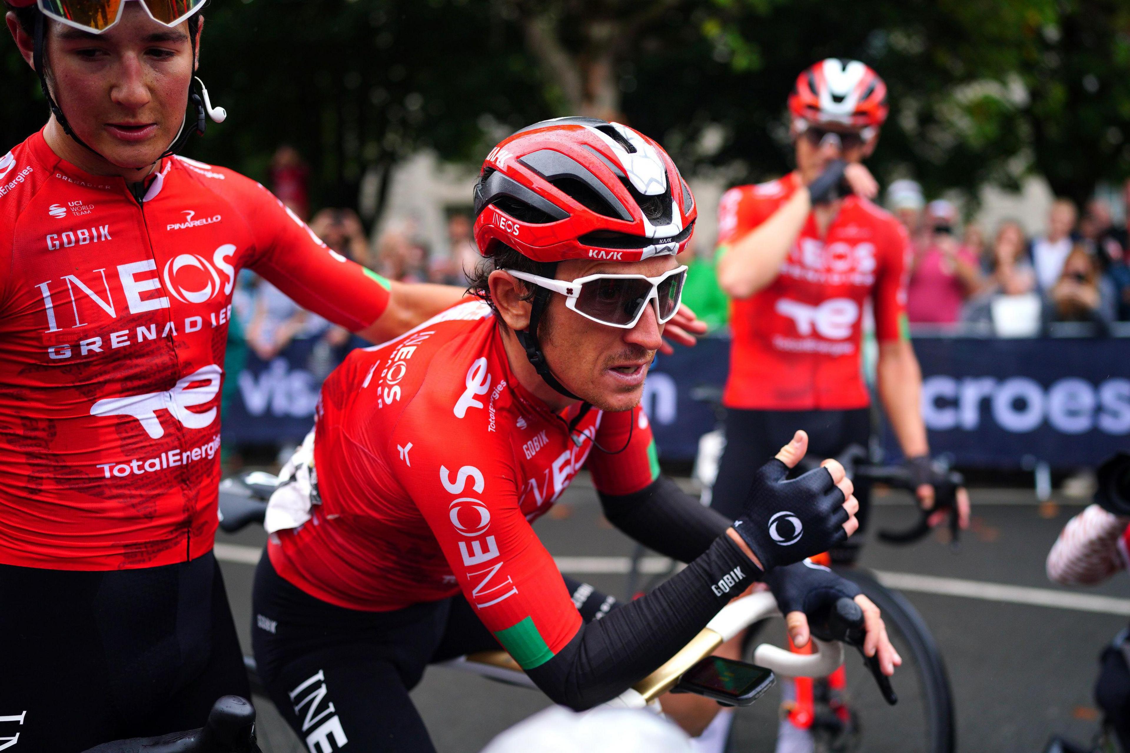 Geraint Thomas after the race from Newport to Cardiff. He has a red jersey on and a red helmet and is on his bike. There is a rider stood to his red in the same red jersey and one to his left too. The crowds can be seen behind him.