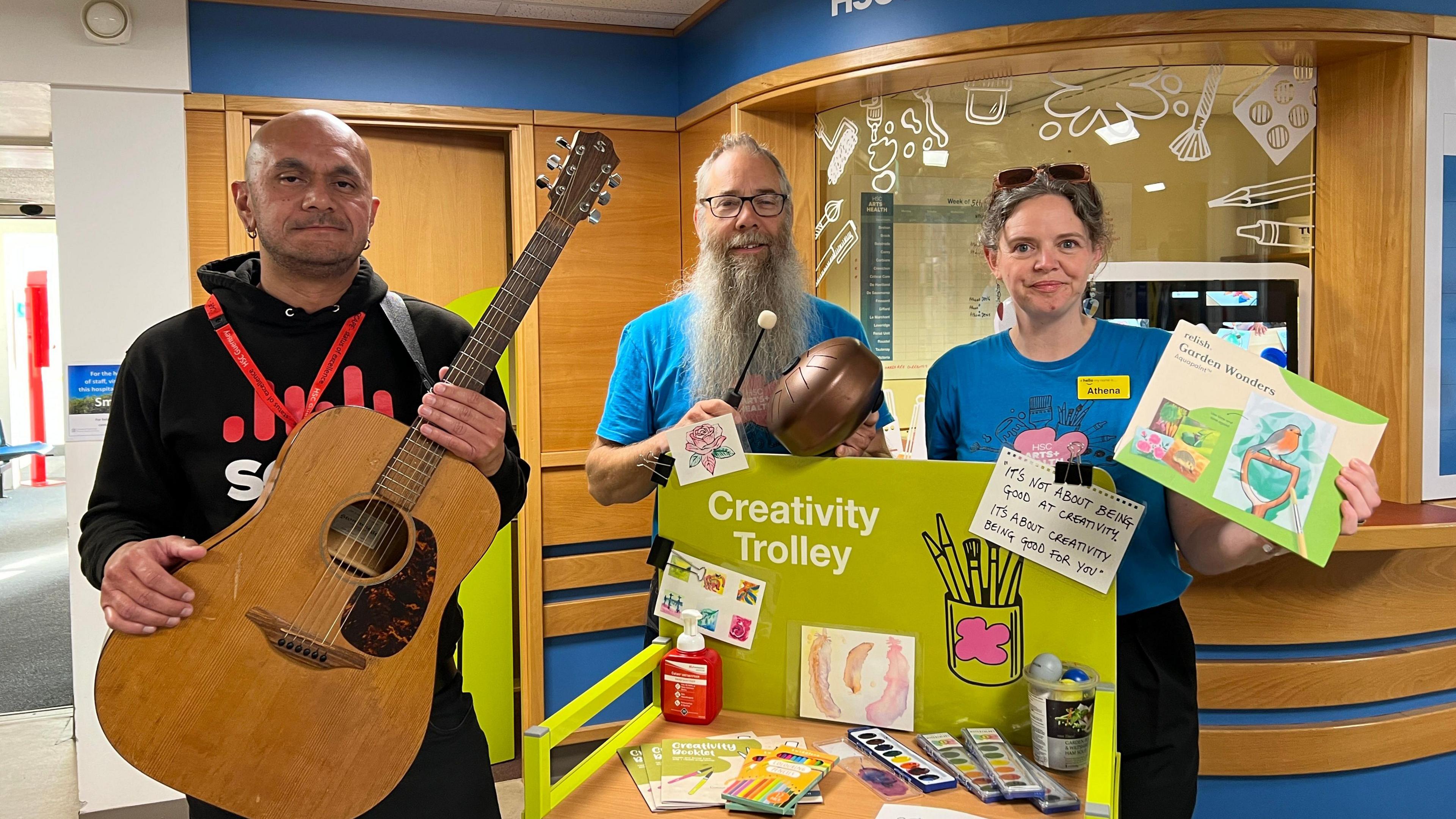 Denis Salgado from the School of Popular Music and Marcel Lenormand and Athene Sholl from Guernsey Art Network stood in the hospital. Denis is holding a guitar, while Marcel and Athene are stood behind a trolly full  or art supplies. 