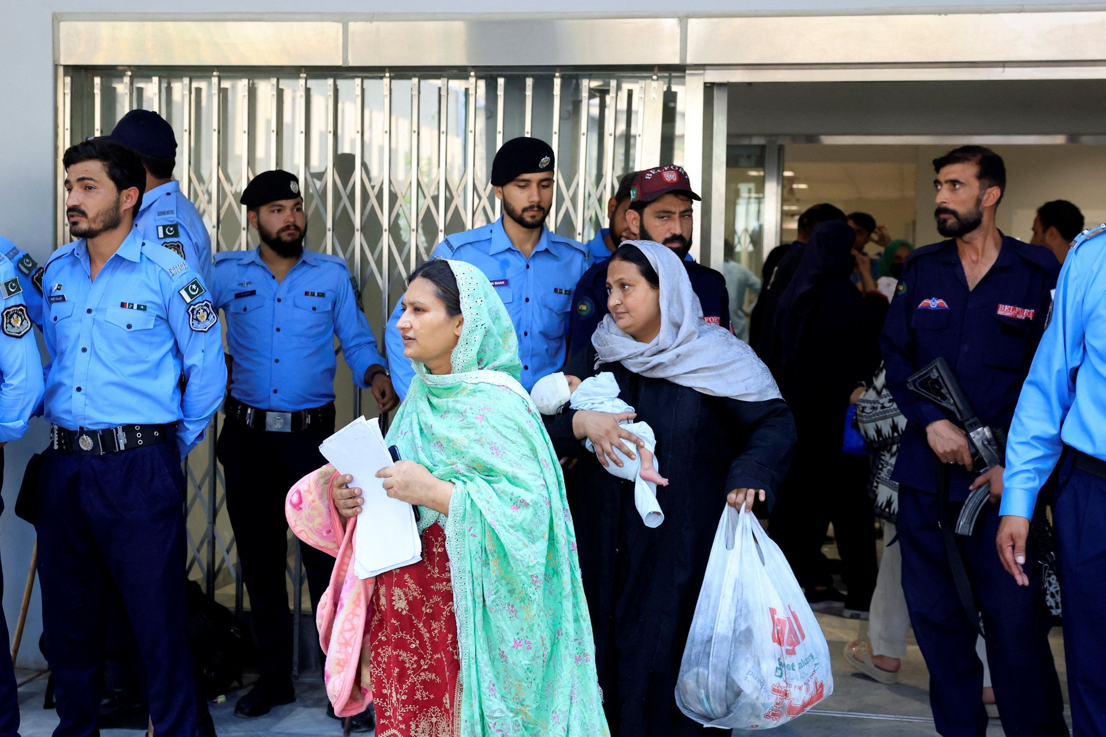 Two women, one carrying a baby, wearing headscarves leave the hospital ward surrounded by police.