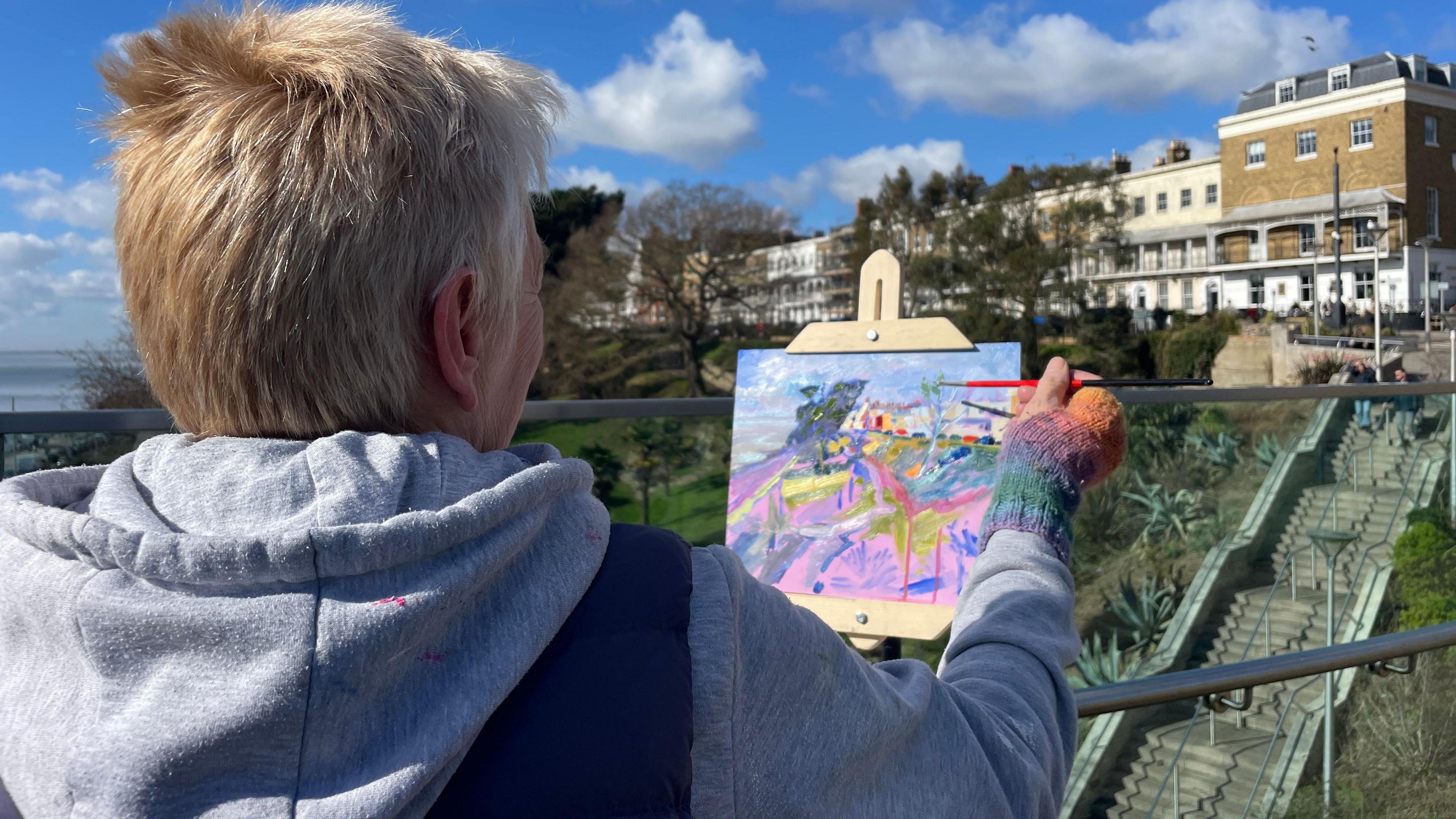 An artist painting a small canvas. The image is colourful and is an interpretation of the view of buildings and park.